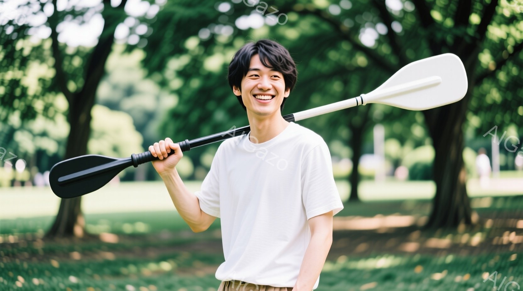 若い男性が白いTシャツとベージュ色のズボンを着用し、笑顔でカヌーのオールを肩に担いで立っている。背景には緑豊かな公園の風景が広がり、木々が生い茂っている。全体的に明るく、自然の中でリラックスした雰囲気を感じさせる写真である。
