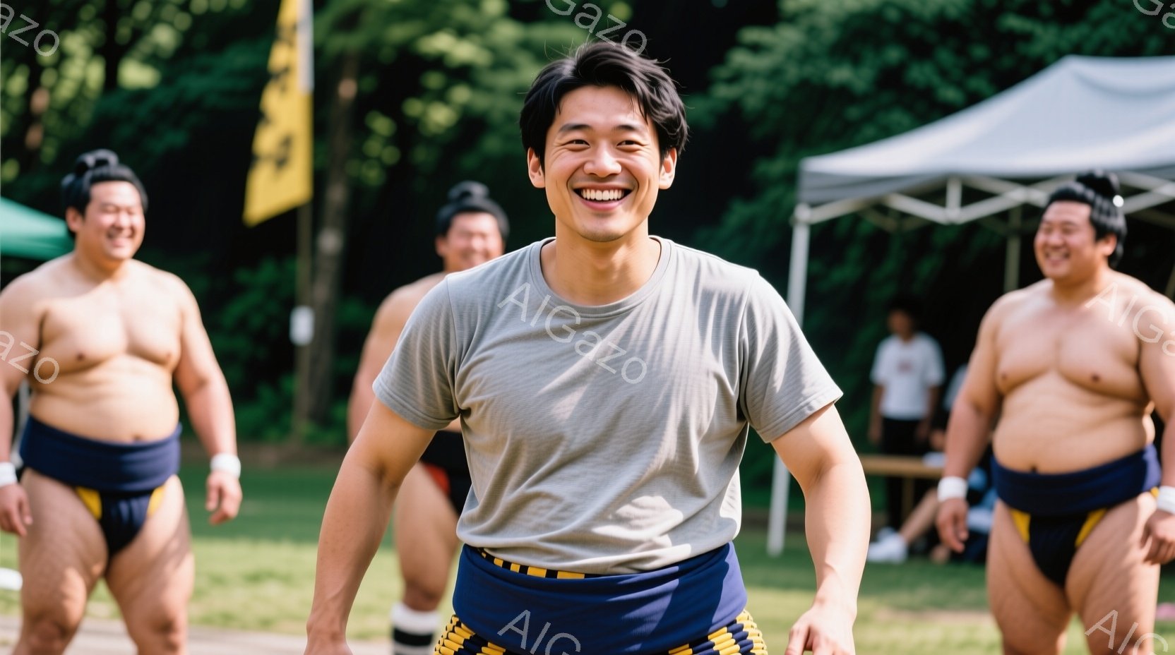 画像中央には、柔道の着（道着のような白いシャツと藍色の締め込み）を身につけた男性が笑顔で立っています。彼の背後には、同じく締め込み姿の太った男性たちが複数おり、彼らもまた笑顔を見せています。背景には緑豊かな木々や白いテントがあり、晴天の下で屋外で行われている柔道の練習風景であることがわかります。