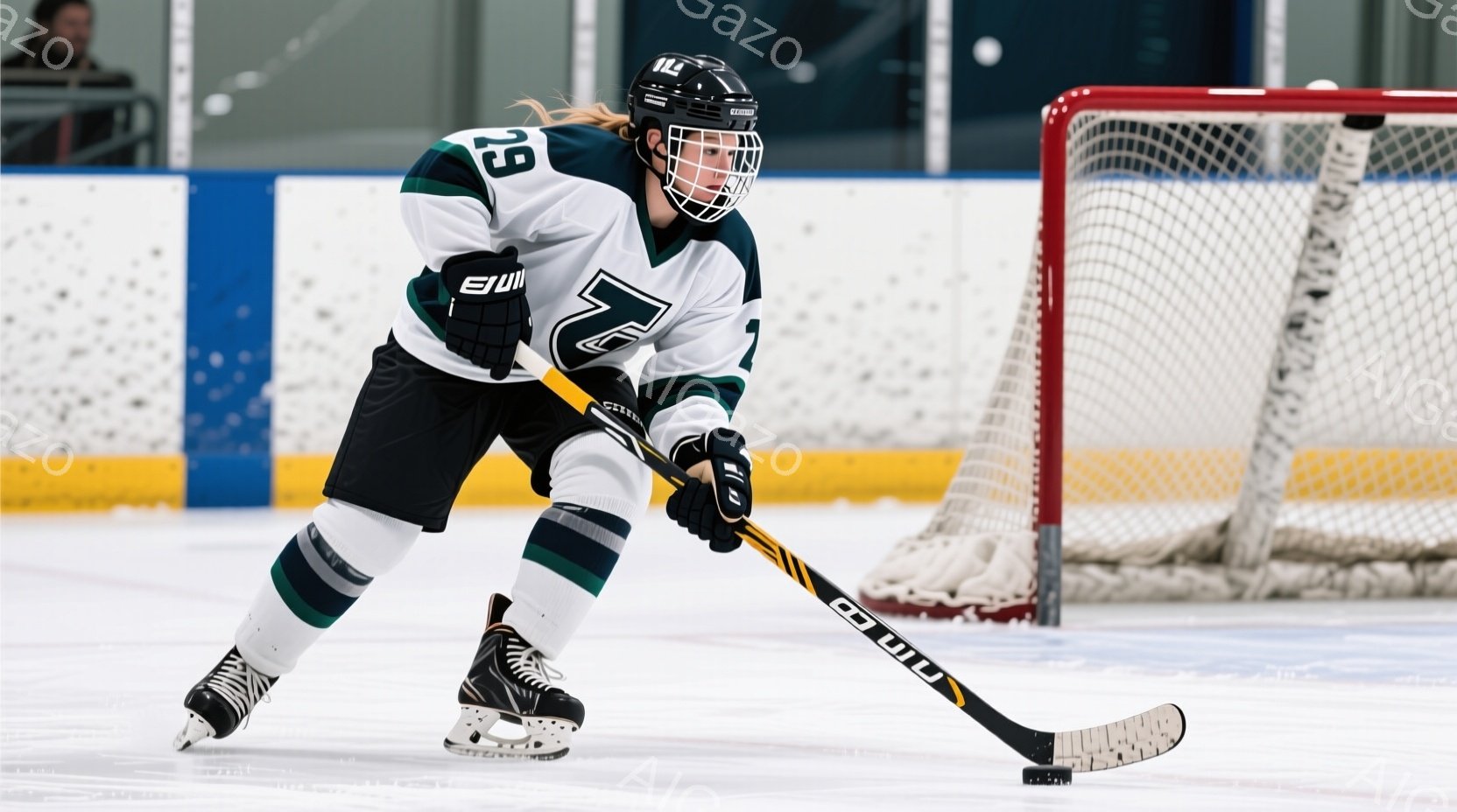 アイスホッケー選手がリンク上に立っており、緑と白のユニフォーム、ヘルメット、そしてスケートを身につけている。選手は少し前かがみになり、スティックを握って集中した表情でいる。背景にはネットと観客席が見え、リンク全体が白く明るい雰囲気である。