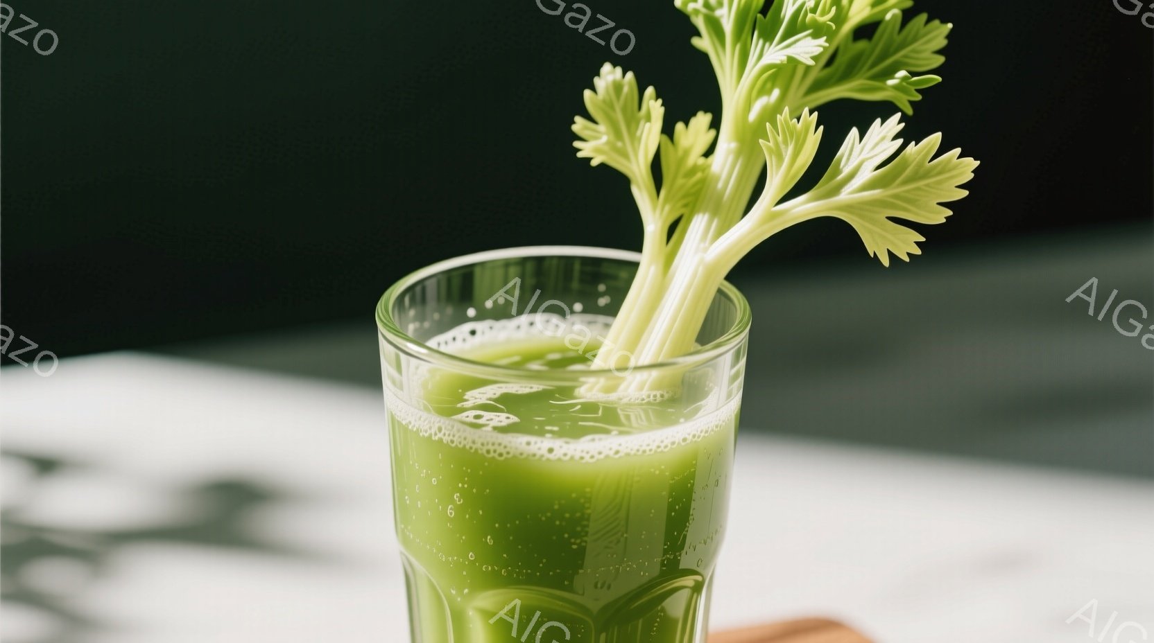 グラスの中に緑色のジュースが入っており、セロリの葉が立てられています。白いテーブルの上にグラスが置かれ、背景には影が落ちています。全体的に健康的で爽やかな雰囲気が漂います。