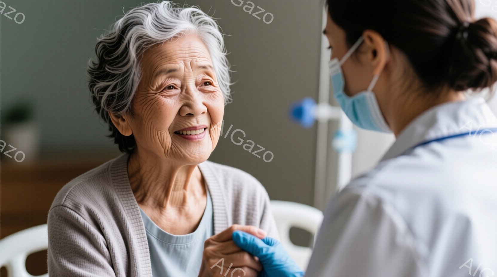 白髪の高齢女性が穏やかな笑顔でカメラを見つめています。彼女は薄いグレーのセーターと水色のインナーを着用し、リラックスした姿勢で座っています。医療従事者と思われる女性が、青い手袋をつけた手で彼女の手を握り、彼女の隣に立っています。