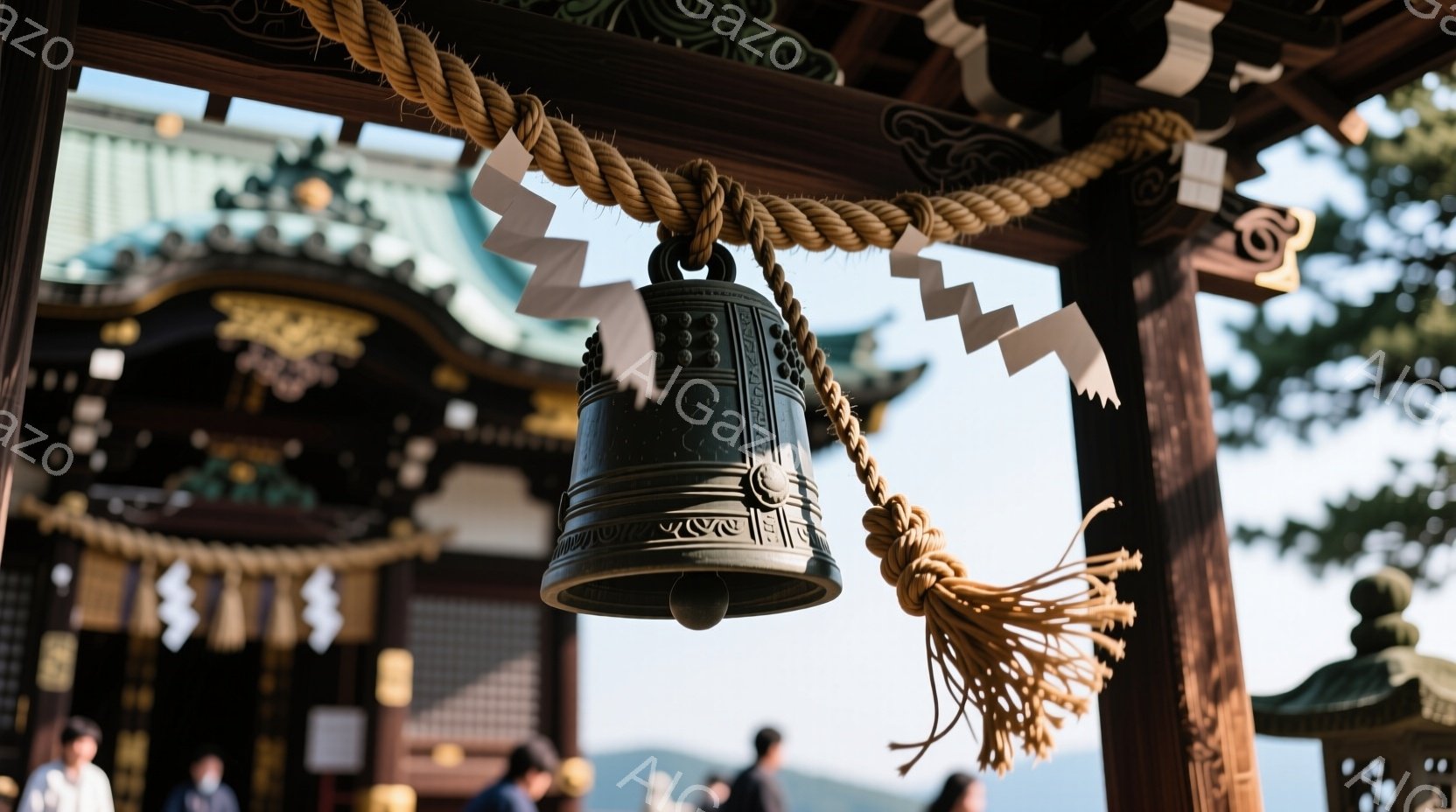 神社の境内にある大きな釣鐘が、木製の柱に吊るされています。鐘の周りには、白い紙の帯が飾られ、下には古びた麻ひもが垂れ下がっています。背景には、鮮やかな朱色の社殿と緑豊かな木々が見え、晴天の下、厳かで静謐な雰囲気が漂っています。