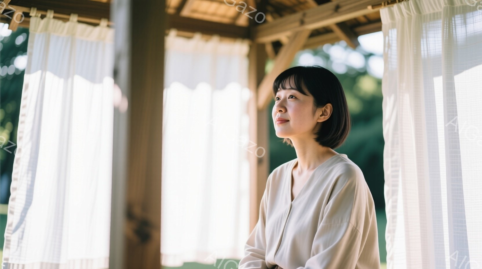 ショートヘアの女性が、薄茶色のシャツを着て、少し上を見ている。彼女はリラックスした姿勢で、背景には木製の柱と白いカーテン、そして緑豊かな自然が見える。全体的に穏やかで、落ち着いた雰囲気の写真である。