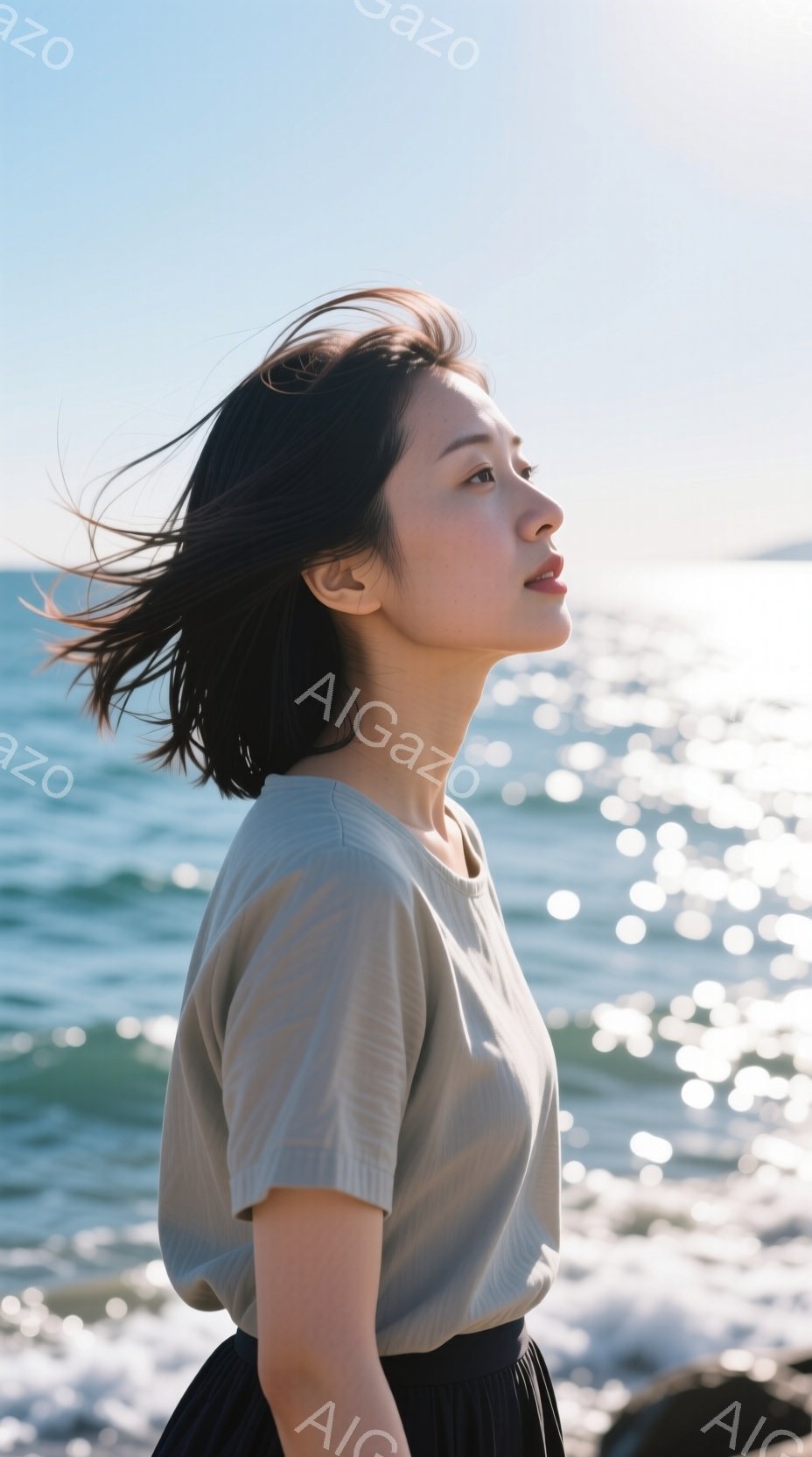 海岸を背景に、横顔の女性が写っている。ベージュ色のシンプルなTシャツと黒いスカートを着用し、風になびく髪が印象的だ。彼女は遠くを見つめ、穏やかで物憂げな表情をしている。