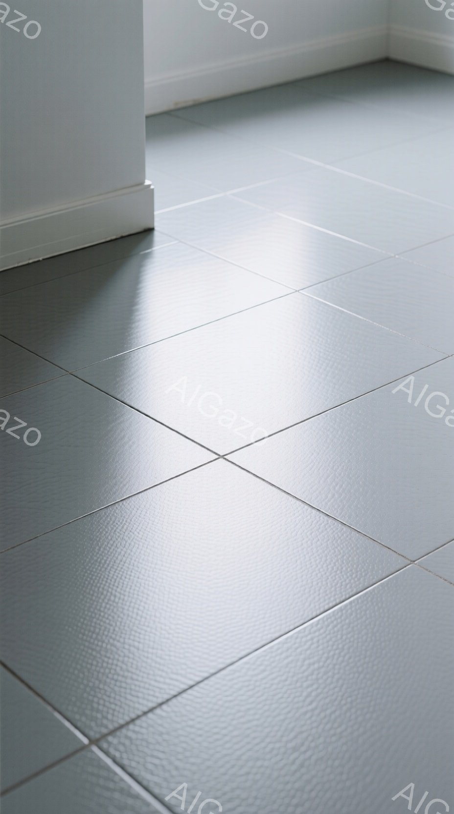 画像は、灰色の正方形のタイルが敷き詰められた床の一部を捉えています。左端には白い壁の一部が見え、タイルと壁の境目に影が落ちています。全体的にシンプルでミニマルな印象で、清潔感と落ち着きを感じさせます。