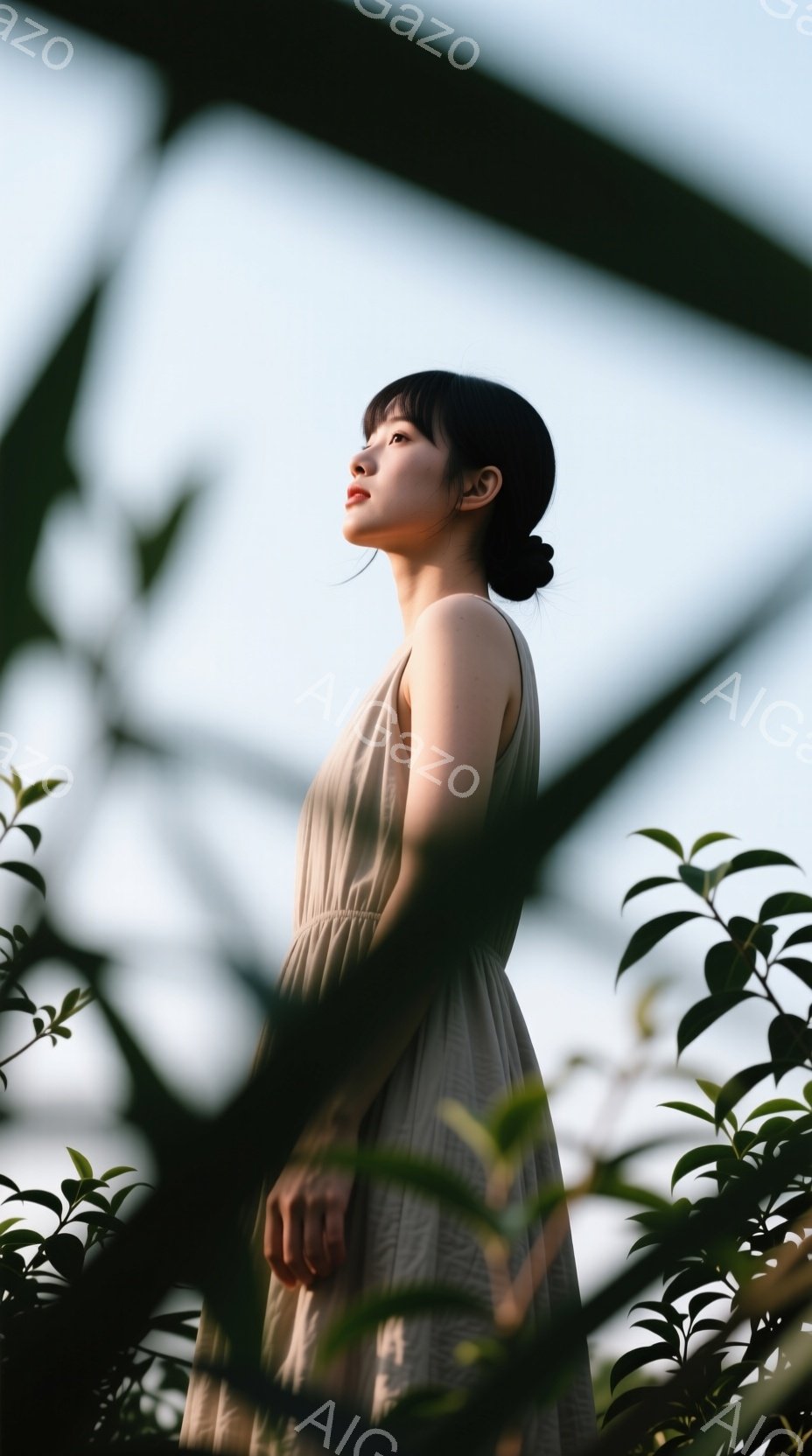 長い髪を後ろで一つにまとめた女性が、薄いベージュ色のワンピースを着て立っている。彼女は横顔で、少し上を見ているように見える表情は穏やかで、柔らかい光に照らされている。背景には緑豊かな植物が見え、その影が女性の周りに落ちて、自然な雰囲気を醸し出している。