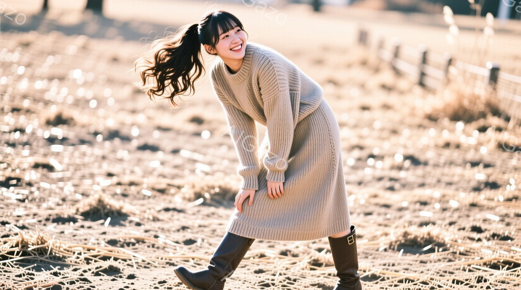 長い茶色の髪をポニーテールにした若い女性が、ベージュのニットワンピースと黒いブーツを着用し、楽しそうに笑っています。彼女は少し体を傾け、まるで踊っているかのように片足を上げています。背景には乾いた草地が広がり、遠くにぼやけた柵が見え、全体的に暖かく穏やかな雰囲気です。