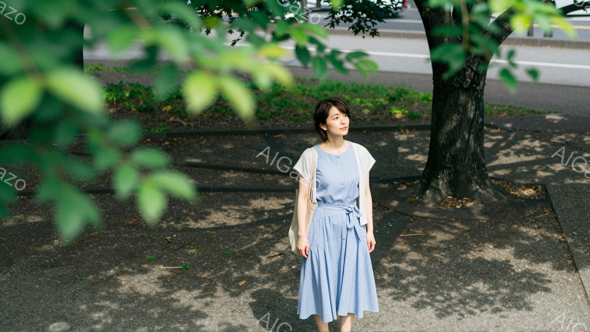 画像には、青いワンピースを着た若い女性が立っています。彼女は肩にバッグをかけ、少し微笑んでいます。背景には木々やアスファルトの道が見え、晴れた日の穏やかな雰囲気が漂っています。