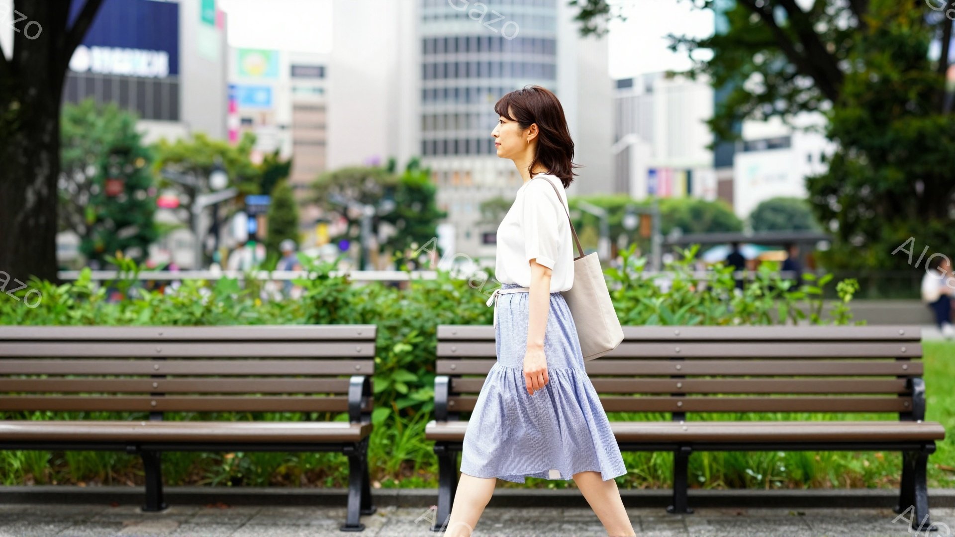 画像には、白いブラウスと水色のプリーツスカートを着用した女性が写っています。彼女はベージュ色のショルダーバッグを持ち、公園の中を歩いています。背景には高層ビルがそびえ立ち、緑豊かな植物と木製のベンチが並び、晴れた日の穏やかな雰囲気を醸し出しています。