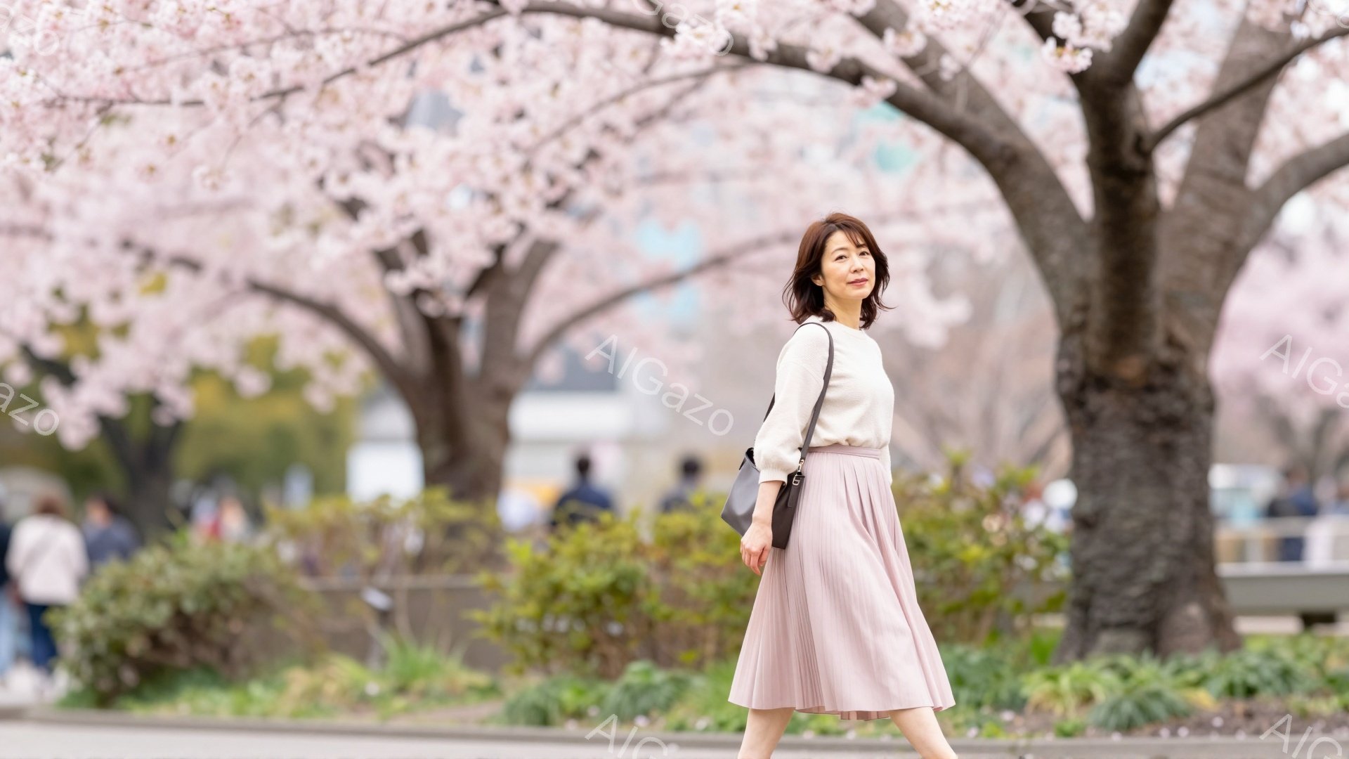 画像には、ピンク色の桜並木を背景に、ベージュのセーターとピンク色のプリーツスカートを着用した女性が歩いている様子が写っています。彼女は肩に黒いバッグをかけ、少し微笑みながら前方を見つめており、春らしい穏やかな雰囲気を醸し出しています。背景には、ぼやけた人々と緑の植生、そして遠くに建物が見え、晴れた日の散歩を楽しんでいる様子が伝わってきます。