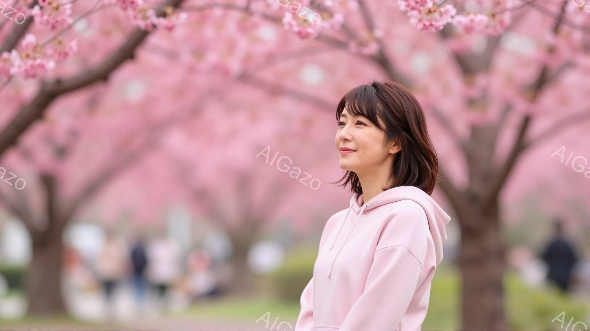 若い女性が桜並木の公園に立っています。彼女はピンク色のパーカーを着ており、少し内側に巻いた茶色の髪が特徴的です。彼女は顔を少し上げて何かを見つめており、穏やかな笑顔を浮かべています。