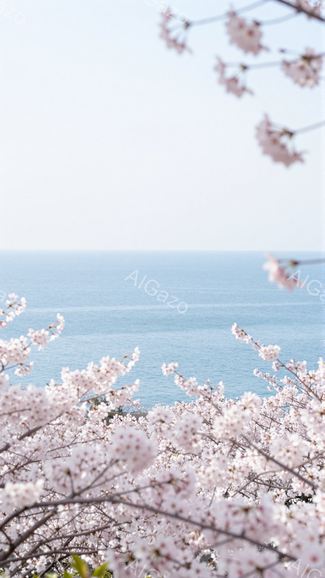 満開の桜の花が手前に咲き誇り、その奥には穏やかな青い海が広がっている。桜の花びらが視界いっぱいに広がり、春の暖かさと優しさを感じさせる美しい風景だ。空は晴れ、海面は穏やかで、全体的に平和で穏やかな雰囲気が漂っている。