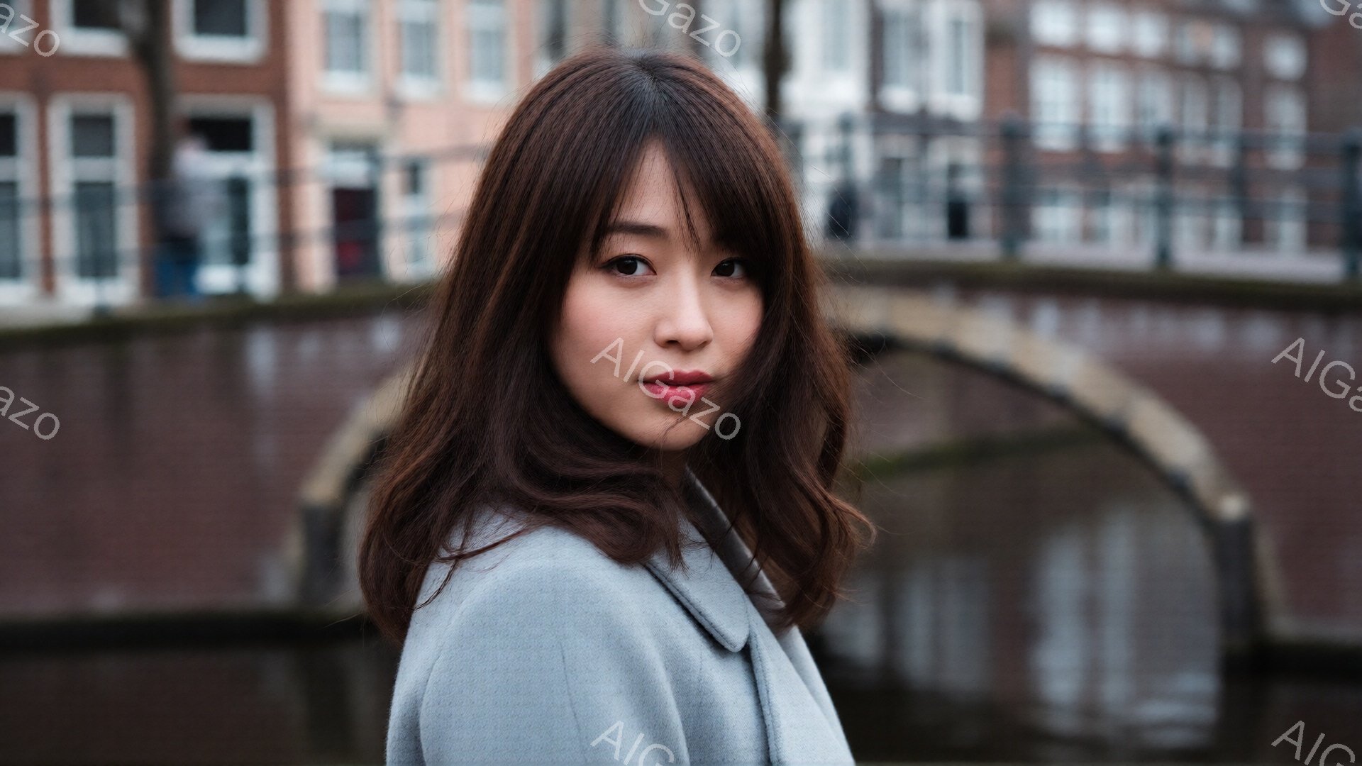 長いダークブラウンの髪を肩にかかるように流した若い女性が写っている。グレーのコートを着ており、少し微笑んだ表情でカメラを見つめている。背景には運河と歴史的な建物が見え、落ち着いた雰囲気の都市の風景が広がっている。