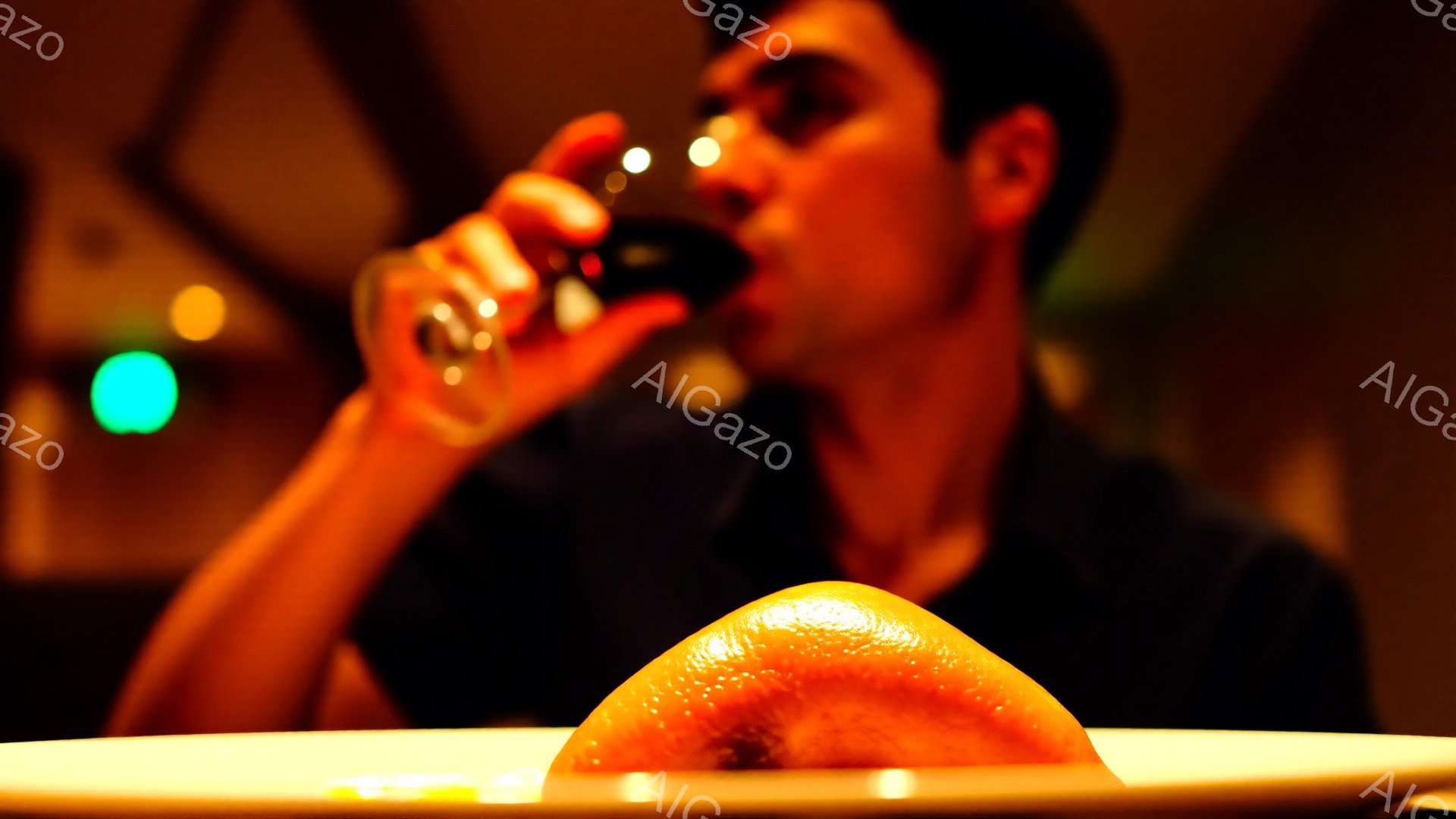 焦点は、白皿に置かれたオレンジ色のゼリーのようなデザートに合わされています。背景には、黒いシャツを着た男性がグラスから何かを飲んでいるぼやけた姿が見え、温かい光が室内を照らしています。全体的に、リラックスした、親密な雰囲気の夕食やカフェのシーンを連想させます。