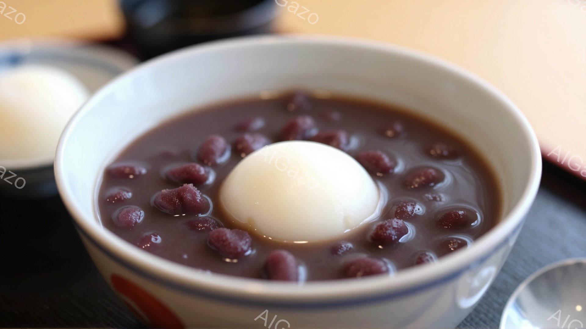 白いお椀に、あんこと白玉が盛り付けられています。あんこは深赤色で、白玉とのコントラストが際立っています。背景には他の器がぼんやりと見え、和やかな食卓の雰囲気が伝わってきます。