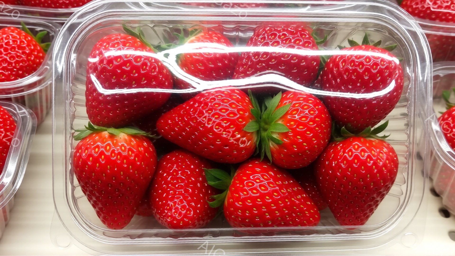 鮮やかな赤いイチゴが透明なプラスチックの容器に並べられています。イチゴはそれぞれ緑色の萼（がく）が付いており、瑞々しく、みずみずしい様子が伝わってきます。背景には同じ容器に入ったイチゴが並んでおり、ス - AI生成フリー素材