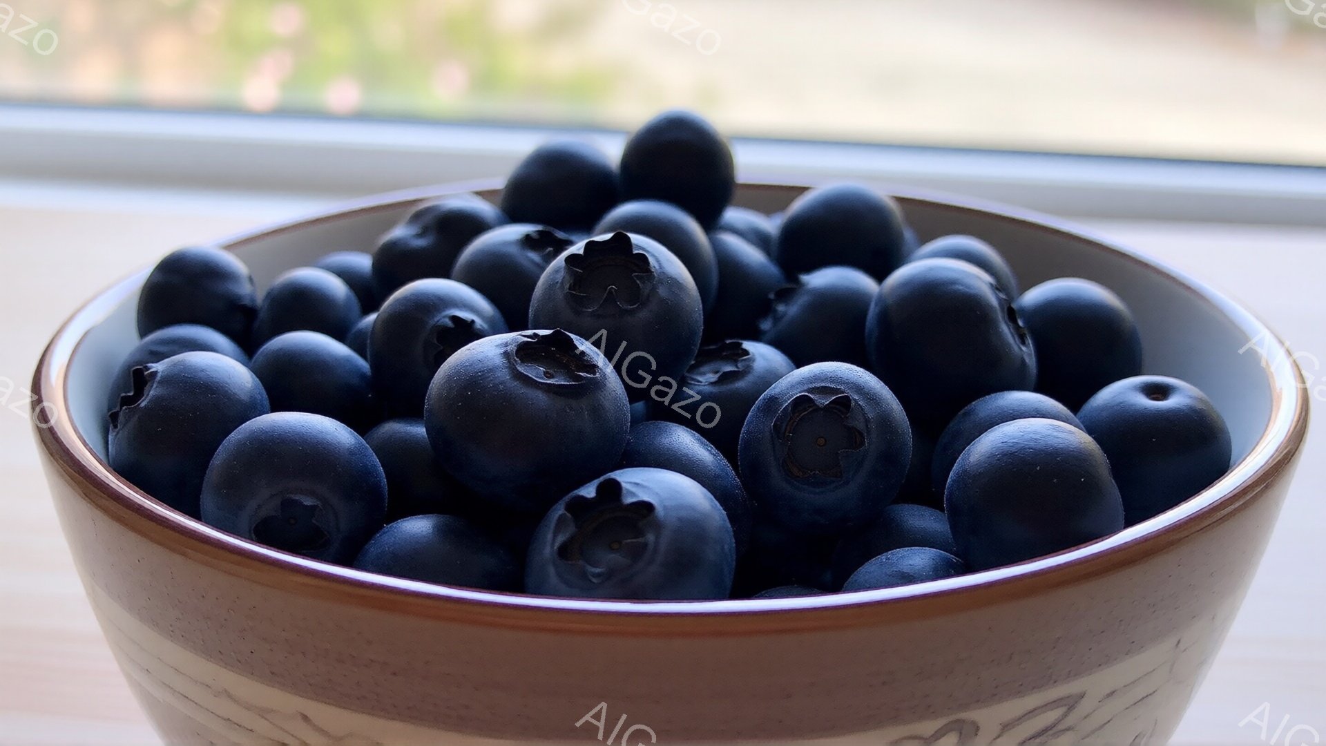 陶器のボウルにたっぷりとブルーベリーが盛り付けられています。光沢があり、丸いブルーベリーは深みのある青紫色をしており、新鮮さを感じさせます。背景は木製のテーブルと窓で、自然光が差し込み、全体的に穏やかで食欲をそそる雰囲気です。