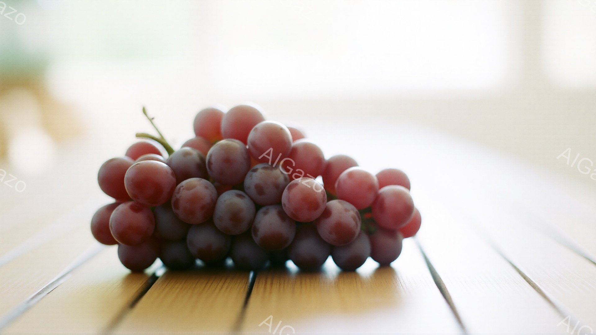 画像には、木製のテーブルの上に置かれた赤紫色のブドウの房が写っています。ブドウは密集しており、表面には白い粉のようなものが付着しているのが見えます。背景はぼやけており、明るい光が差し込み、全体的に穏やかで自然な雰囲気を醸し出しています。