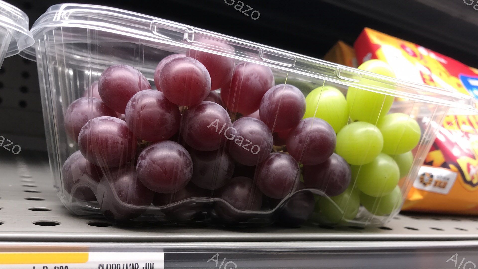 透明なプラスチックの容器に、紫色のブドウと緑色のブドウが並んで入っています。容器はスーパーマーケットの棚に置かれており、背景には他の商品のパッケージが見えます。全体的に、鮮やかで食欲をそそる雰囲気です。