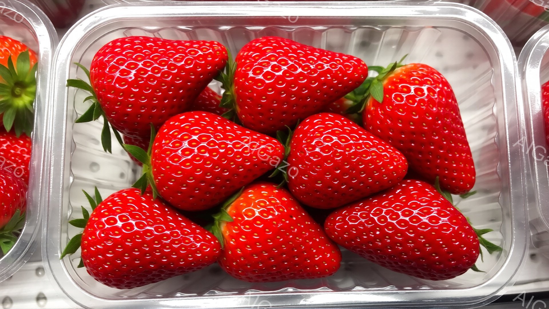 鮮やかな赤色のいちごが透明なプラスチックの容器にぎっしりと詰まっています。いちごはみずみずしく、表面には小さな種が規則的に並んでいます。容器の背景は光沢のある金属で、全体的に明るく清潔な印象を与えます。