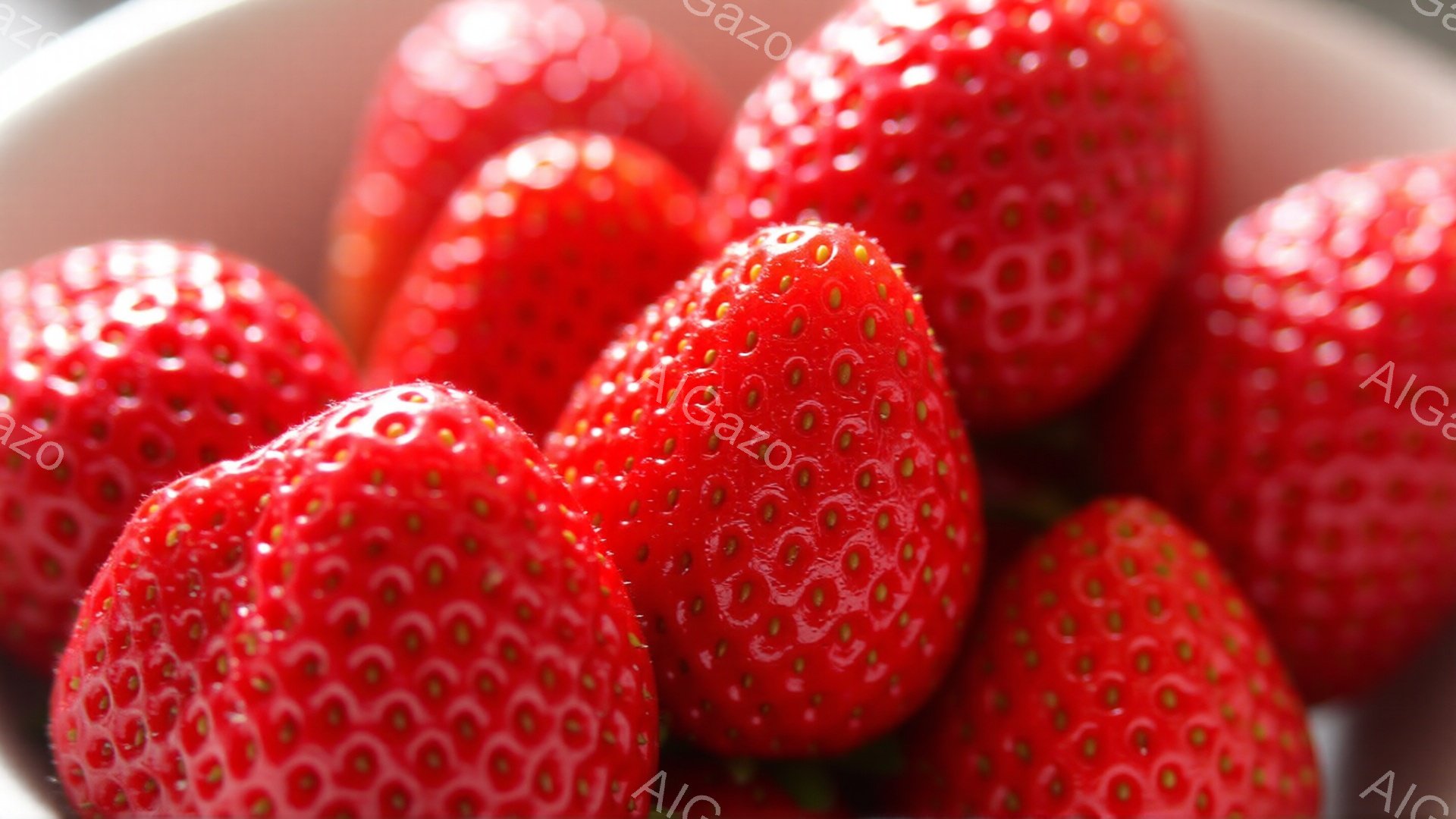 鮮やかな赤色のイチゴが、白い容器の中に密集して写っています。表面には種がびっしりと並び、光沢があり、みずみずしい様子が伝わってきます。背景は白くぼやけており、イチゴの鮮やかさを際立たせています。