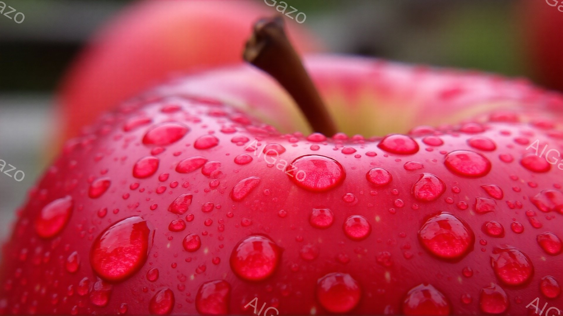 真っ赤なリンゴが、表面に水滴をまとって写っているクローズアップ写真です。リンゴの鮮やかな赤色と丸みを帯びた水滴が、瑞々しさを強調しています。背景は緑色でぼやけており、リンゴが際立って見えます。