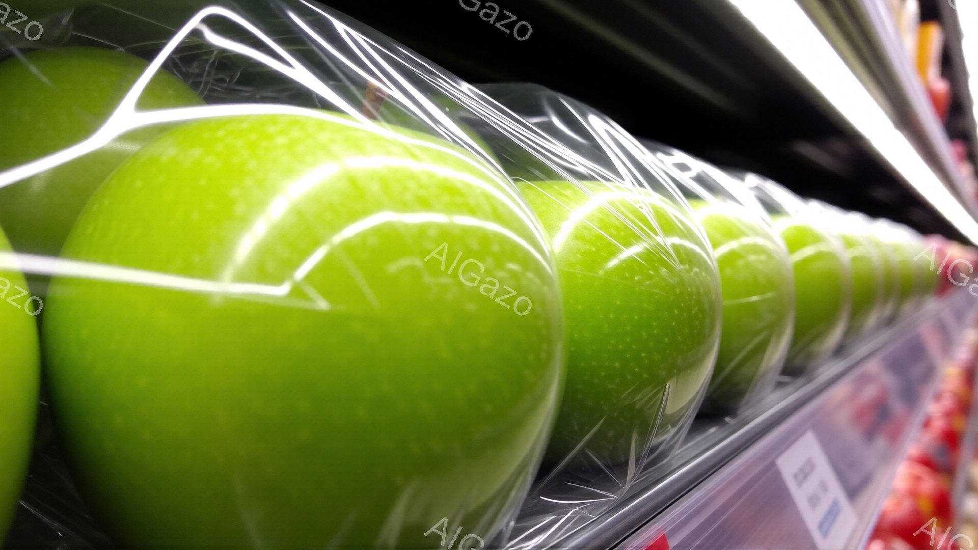 画像には、透明なプラスチックの包装に並べられた鮮やかな緑色のリンゴが写っています。リンゴは均一な大きさで、光沢があり、新鮮に見えます。背景はぼやけており、店舗の棚の一部が見え、全体的に清潔で整理された印象を与えます。