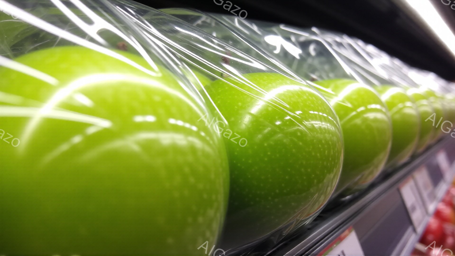 鮮やかな緑色のリンゴが透明なプラスチックのケースに並べられています。リンゴの表面には光が反射し、つややかな質感が際立っています。背景には赤い果物が見え、スーパーマーケットか八百屋のような小売店の棚の一部であると思われます。