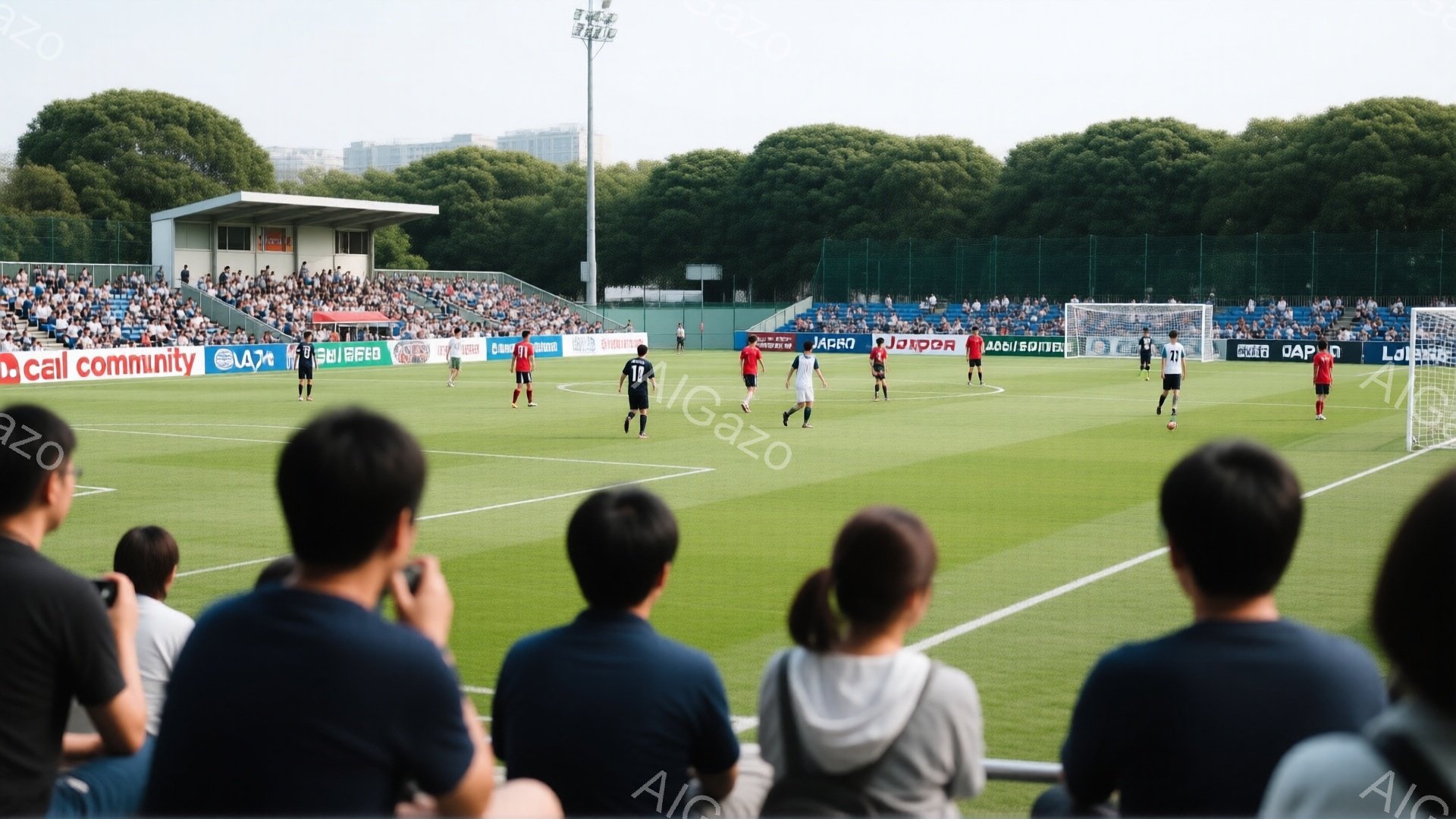 サッカーの試合が行われている様子が写っている。観客席からは複数の観客がフィールドを見下ろしており、何人かはスマートフォンで写真を撮っている。背景には緑豊かな芝生、観客席、そして遠くの建物や木々が見え、晴れた日の屋外でのスポーツイベントの活気ある雰囲気が伝わってくる。