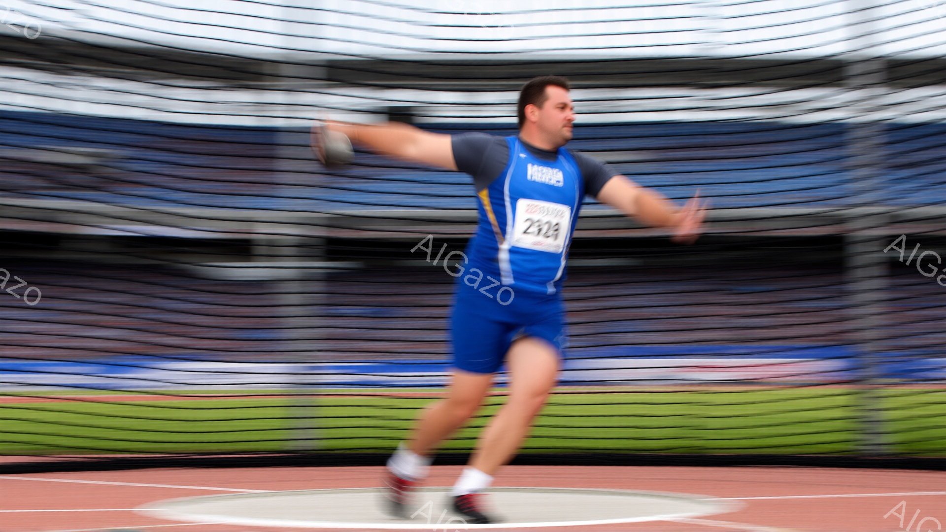 競技者は青いユニフォーム姿で、ディスカスを投げている最中です。彼の姿勢は力強く、飛翔するような動きが捉えられています。背景には観客席が広がり、競技場のトラックが確認でき、天候は晴れで競技に最適な状況です。