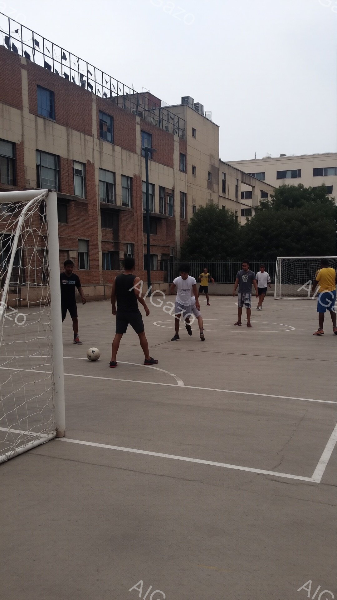 複数の男性が屋外のコンクリートコートでサッカーをしている様子が写っている。彼らは皆、短パンとTシャツ姿で、活発にボールを追いかけている。背景にはレンガ造りの建物が立ち並び、天気は曇りで、コート全体に影が落ちている。