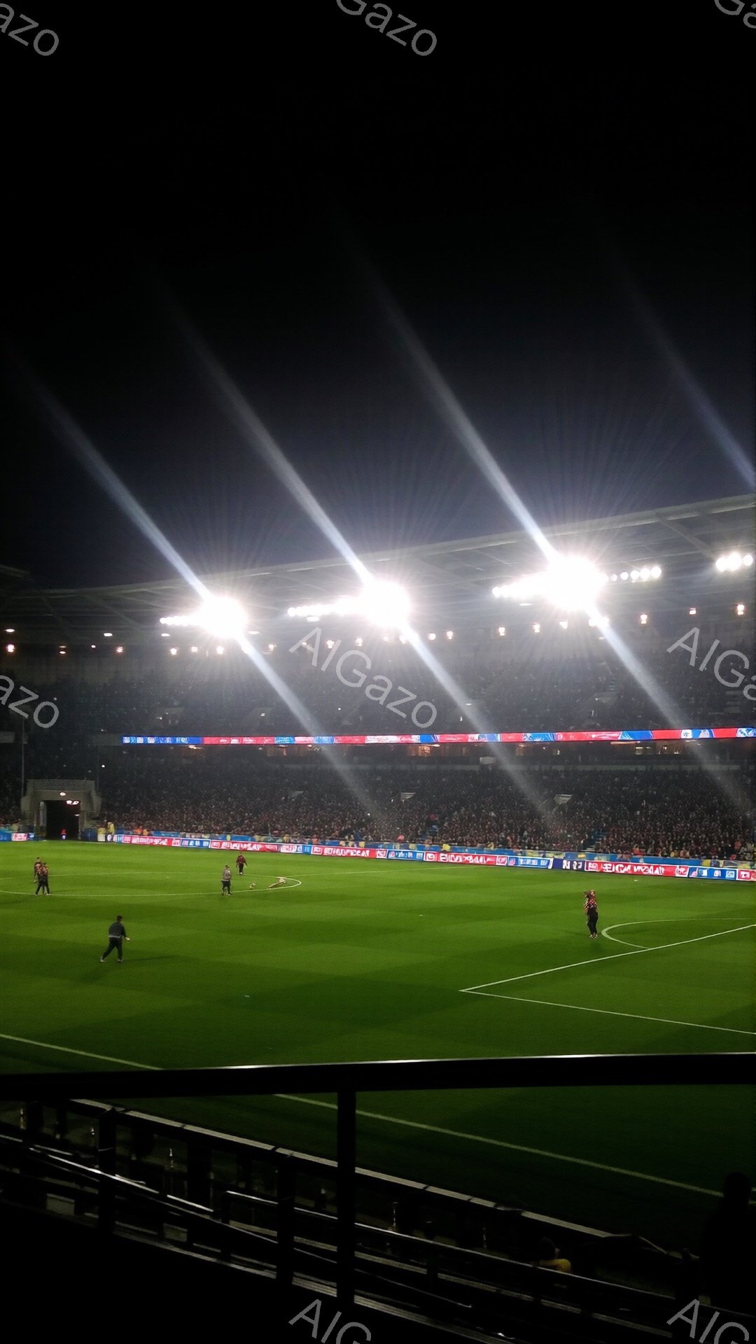 夜間のサッカー競技場は、鮮やかな緑色の芝生と、観客席を埋め尽くす人々の姿が印象的です。フィールド上の選手たちは、赤いユニフォームを着ており、試合に集中して動き回っています。強力なライトが照らし出す芝生と、熱狂的な観客の姿から、激しい試合の雰囲気が伝わってきます。