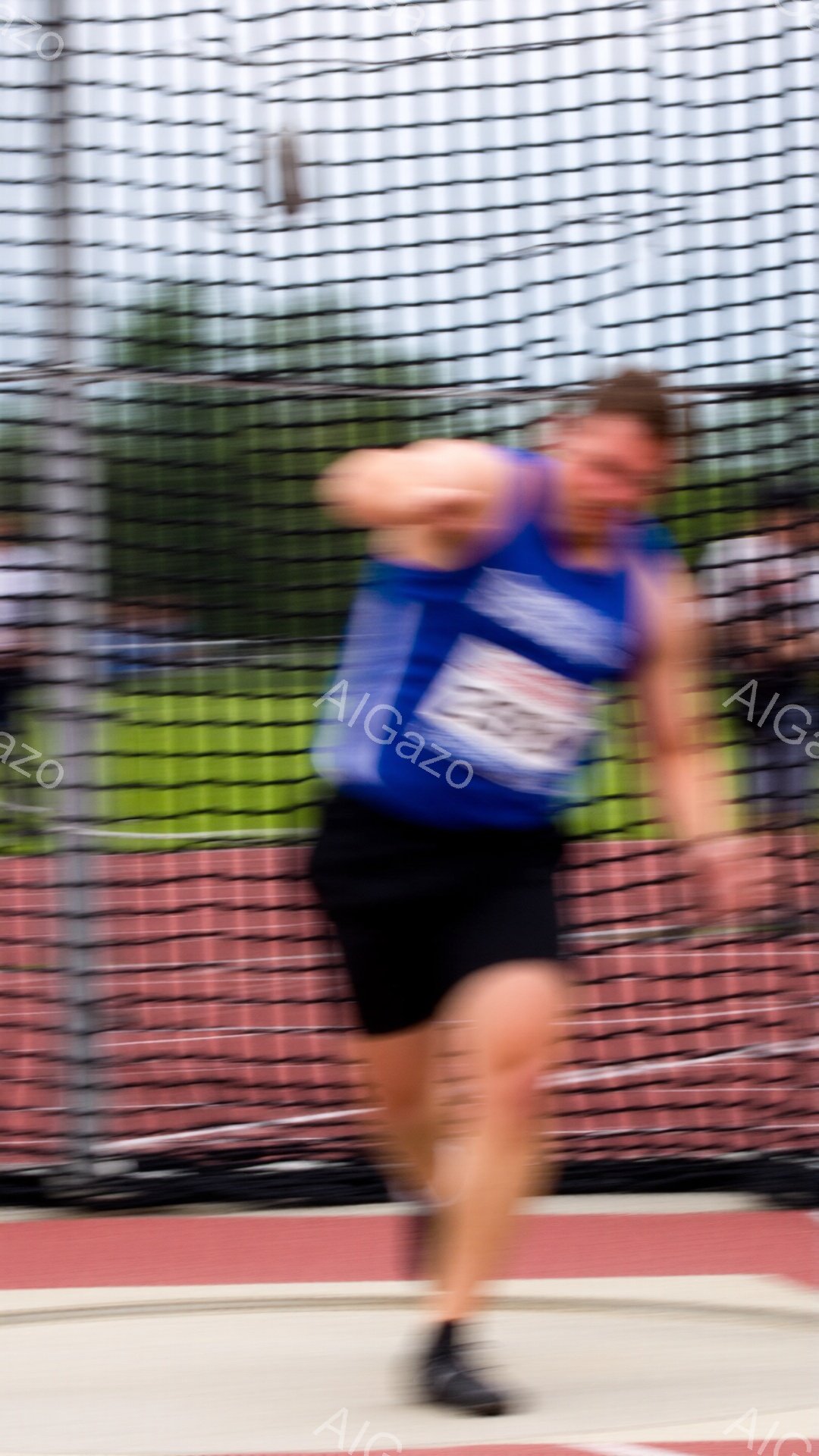 競技者が円盤投げの最中に捉えられた写真で、青いタンクトップと黒い短パンを着用し、強い力で円盤を投げている様子がわかる。背景には赤いトラックと緑の自然が広がり、円盤投げのネットが競技者の動きを捉え、躍動感あふれる雰囲気を醸し出している。競技者の表情ははっきりと見えないが、身体全体から力強さが伝わってくる。