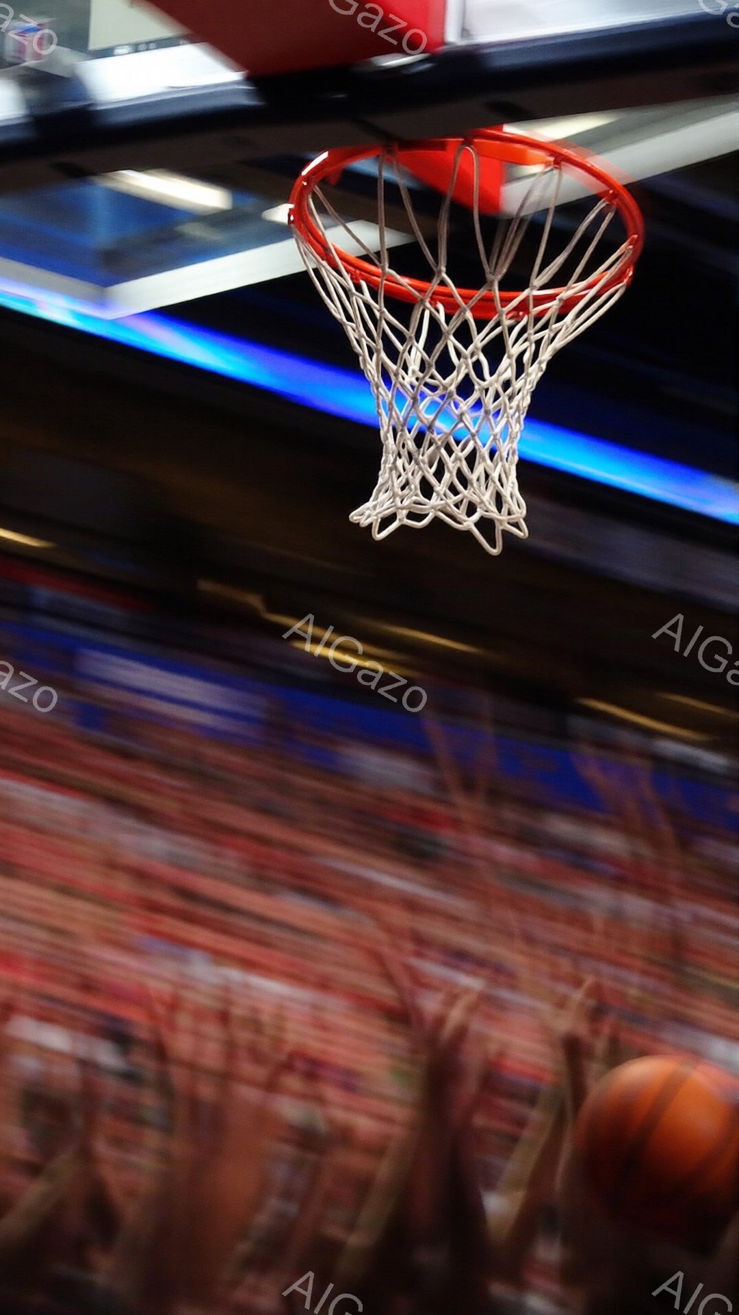 この画像は、バスケットボールのゴールと、激しい動きの中でぼやけた観客席と選手の一部を示しています。ゴールネットは鮮明ですが、背景はモーションブラーにより色彩の帯とぼやけた人影が混ざり合い、試合のスピード感と興奮を強調しています。オレンジ色のバスケットボールが画面の右下に見え、試合の進行を暗示しています。
