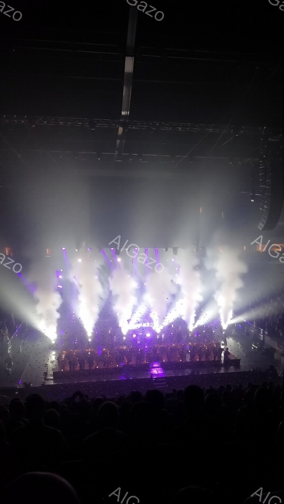 ステージ上には多くの人々が立ち並び、紫と白の照明と紙吹雪に包まれています。彼らは暗い背景の中でシルエットとしてしか見えず、興奮と熱狂的な雰囲気が伝わってきます。会場全体は薄暗く、観客席の一部が見え、コンサートやイベントの最中であることが伺えます。