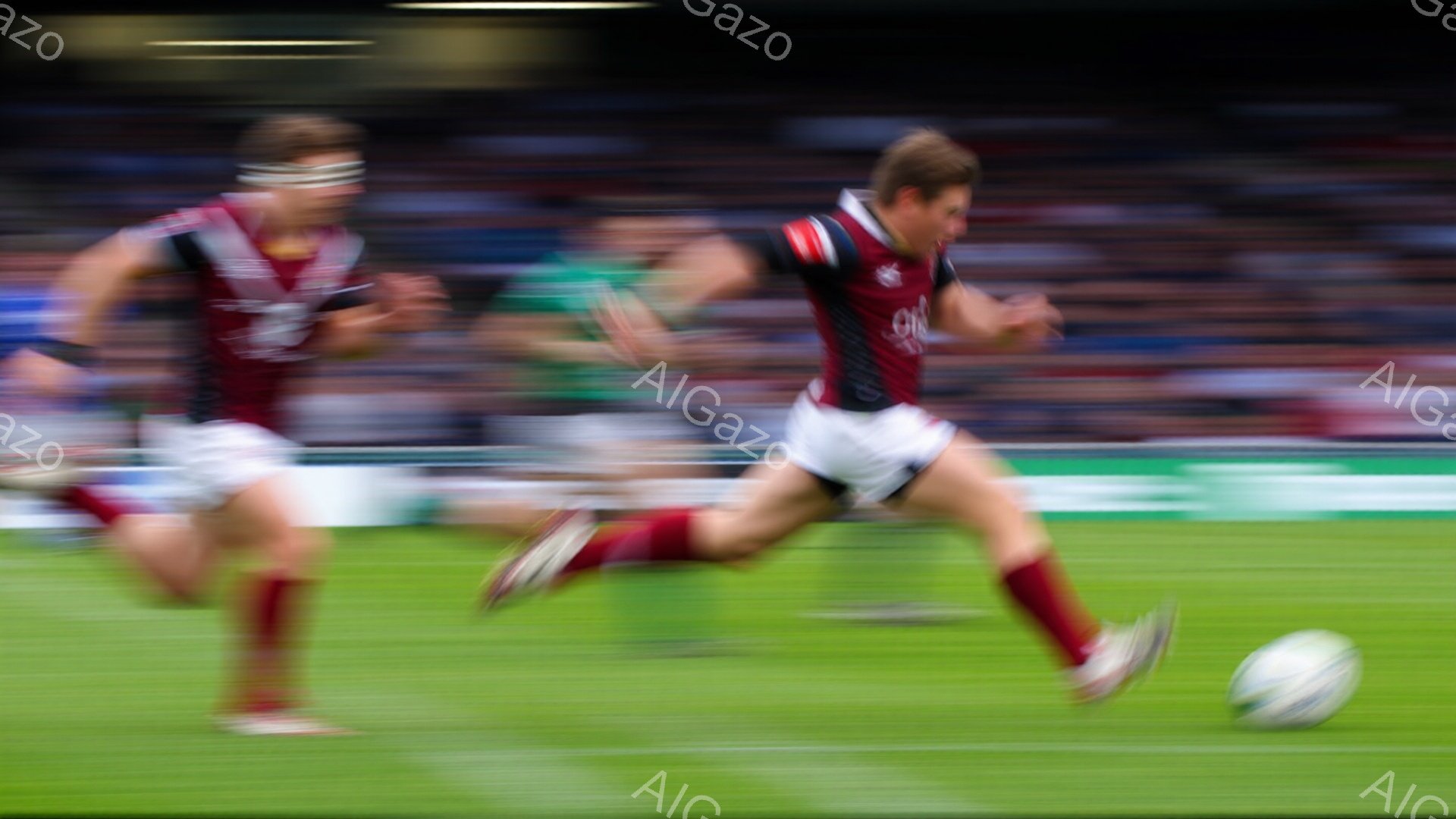 ラグビー選手が緑の芝生の上を全速力で走っている様子が捉えられています。選手は濃い赤色のユニフォームと白いショーツ、赤いソックスを着用しており、髪は短く整えられています。背景にはぼやけた観客席とスタジアムの構造が見え、試合の熱気が伝わってきます。