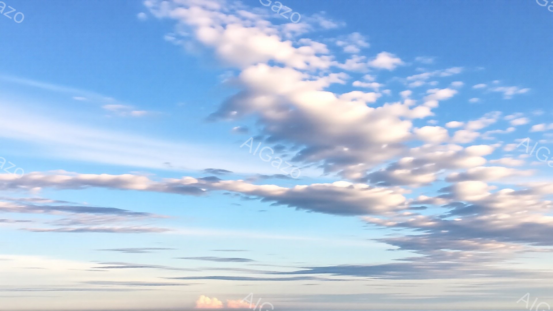 空は明るい水色から淡いピンク色へとグラデーションがかかっており、所々に白い雲が浮かんでいる。雲はもくもくと高く積み重なり、まるで綿菓子のように柔らかい印象を与える。地平線付近には、薄っすらと赤みがかった山並みがぼんやりと見える、穏やかで幻想的な空模様だ。