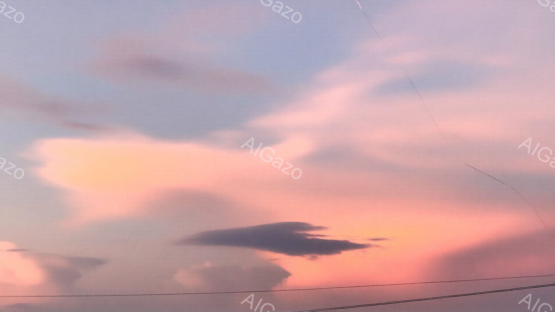 空は夕焼けの名残で、淡いピンクとオレンジ色が広がっている。特徴的なのは、まるで巨大な波のような形をした雲で、空に浮かび、幻想的な雰囲気を醸し出している。電線が画面下部にわずかに見え、日常の風景の中に非日常的な空が広がっている様子が印象的だ。