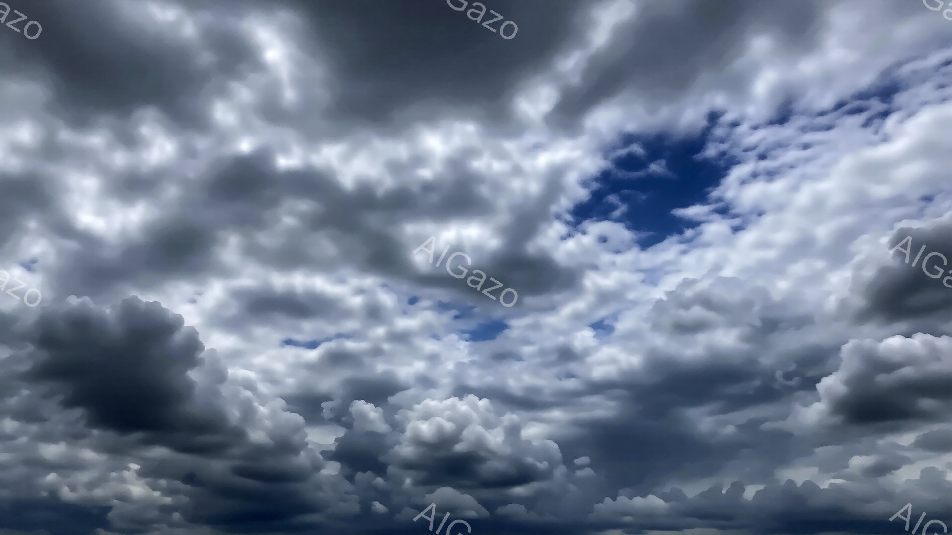 厚い雲が空を覆っており、青空が雲の隙間から覗いています。雲は白とグレーの様々な色合いで、重厚感と動きを感じさせます。これは嵐が近づいている、または過ぎ去った後の空の様子を示唆しています。
