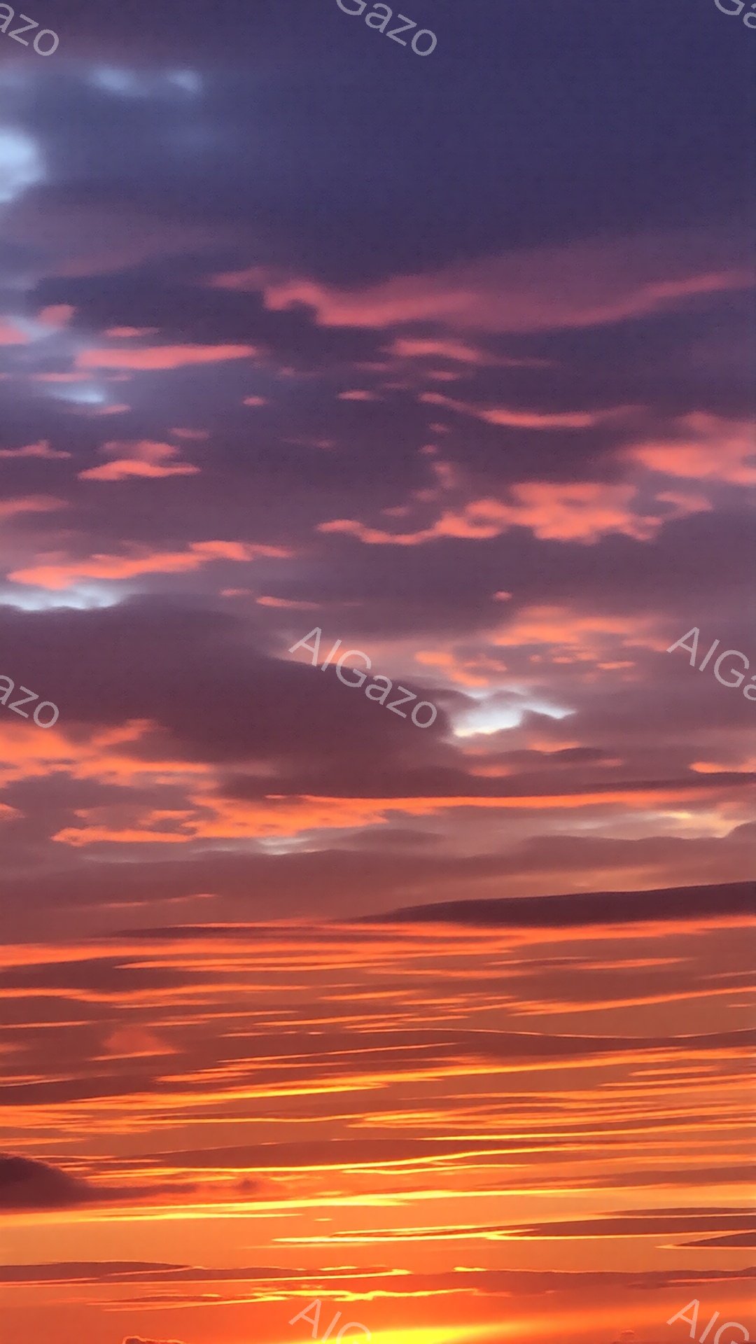 空は燃えるようなオレンジ色と赤色のグラデーションで覆われ、まるで地平線が炎に包まれているようだ。雲は水平方向に長く伸びており、夕焼けの光を反射して金色に輝いている。全体的に、力強く、ドラマチックな、そして壮大な自然の美しさを感じさせる雰囲気だ。