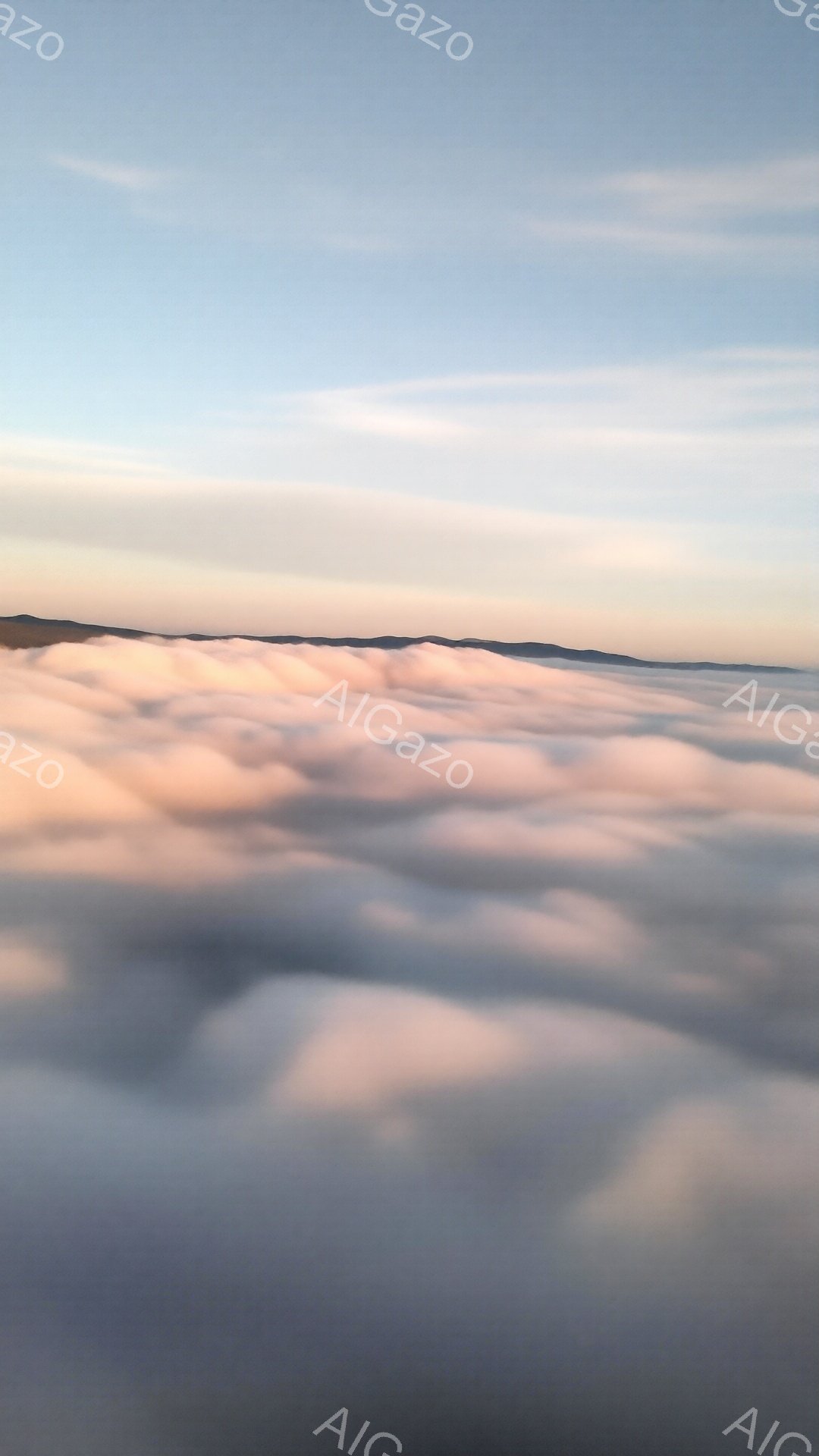空から見た雲海が広がり、朝焼けの柔らかなピンク色が雲を照らしている。雲の海はまるで起伏のある雪景色のように見え、遠くにはぼんやりとした山の稜線が確認できる。全体的に穏やかで幻想的な雰囲気が漂っている。