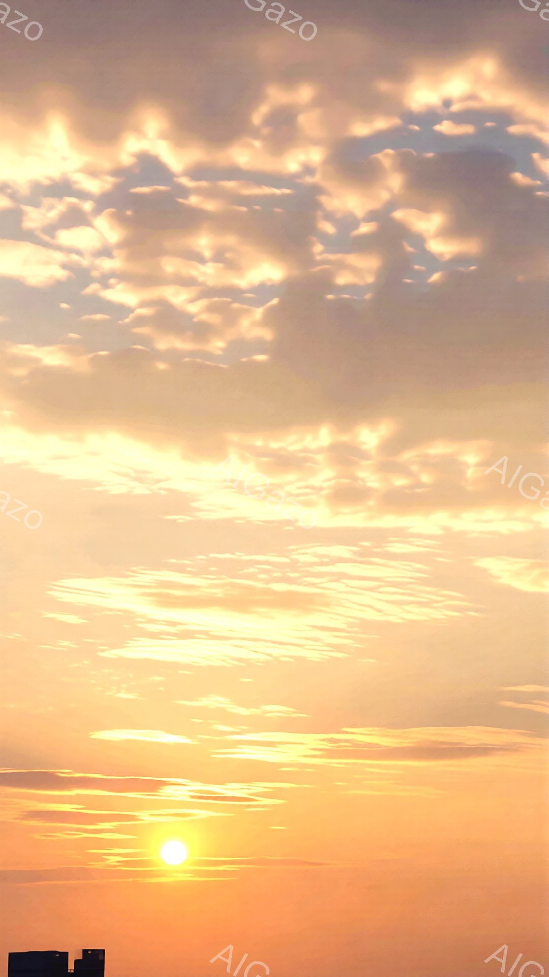 空は夕焼けの色で染まり、オレンジと黄色のグラデーションが広がっています。太陽は地平線近くにあり、強い光を放ち、雲がその周りを縁取るように配置されています。下には建物のシルエットがわずかに見え、穏やかで - AI生成フリー素材