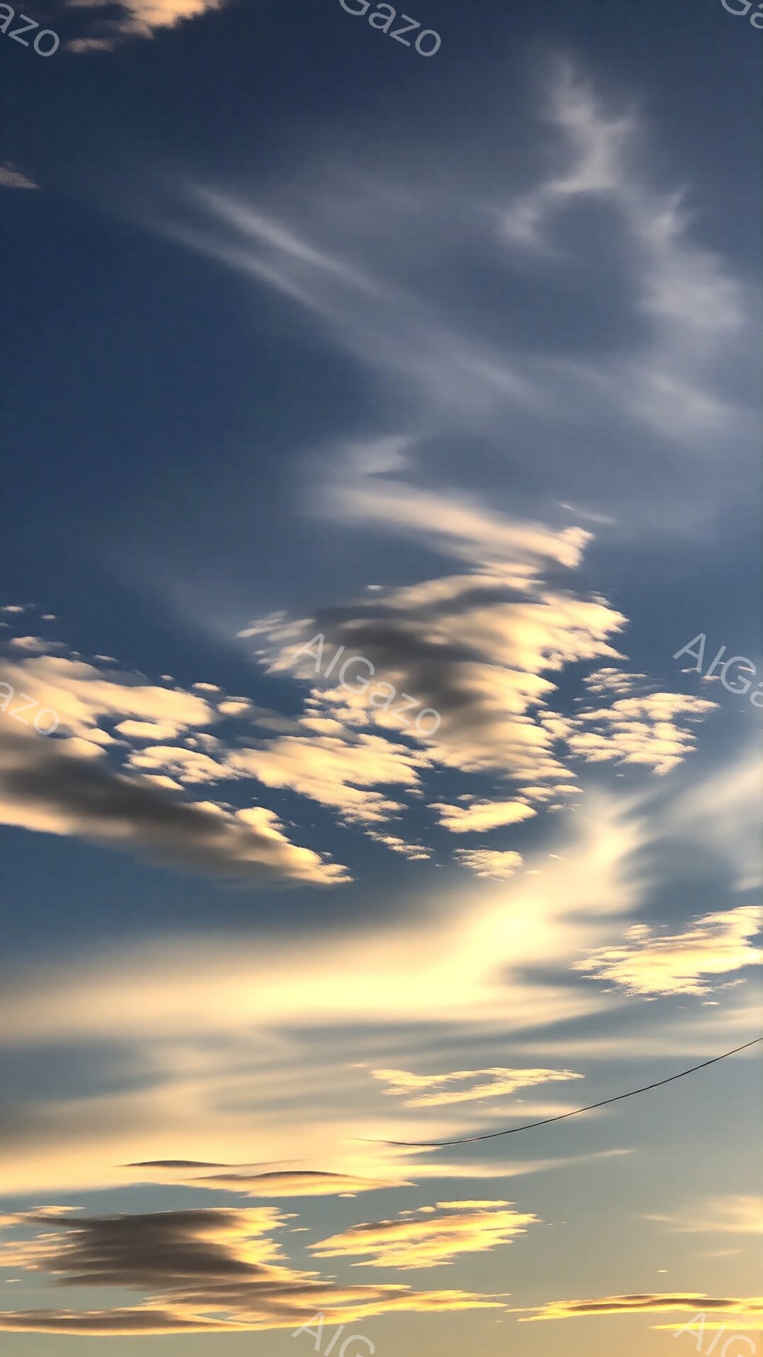 空は鮮やかな青色で、太陽の光を浴びたふわふわとした雲が黄金色に輝いている。雲は様々な形をしており、まるで空に描かれた絵画のようだ。遠くには電線が一本見え、自然と人工物が共存する穏やかな風景を作り出している。