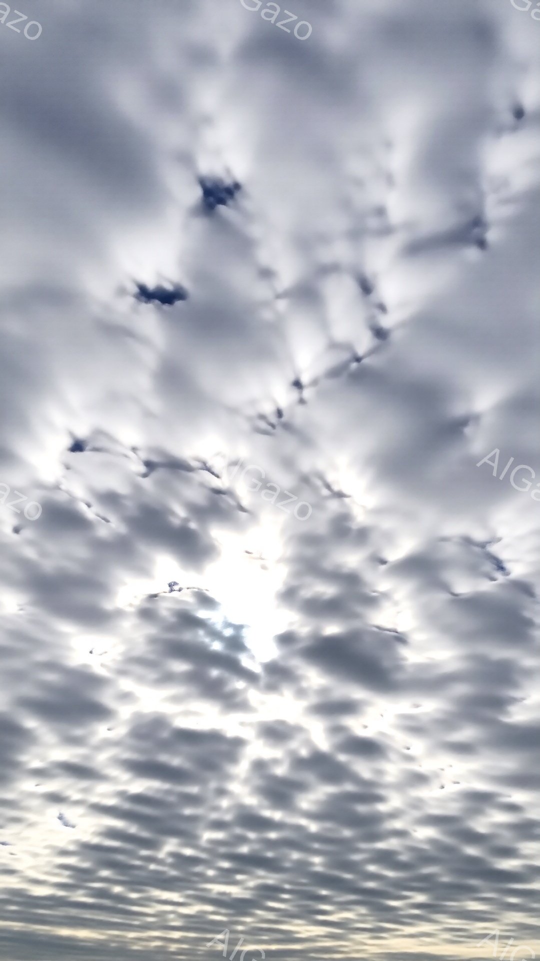 空は細かい雲で覆われ、太陽が雲間から眩しく輝いている。雲はまるで鱗のように重なり合い、規則的な模様を作り出している。全体的に穏やかで静かな雰囲気だが、太陽の強い光がどこか神秘的な印象を与える。