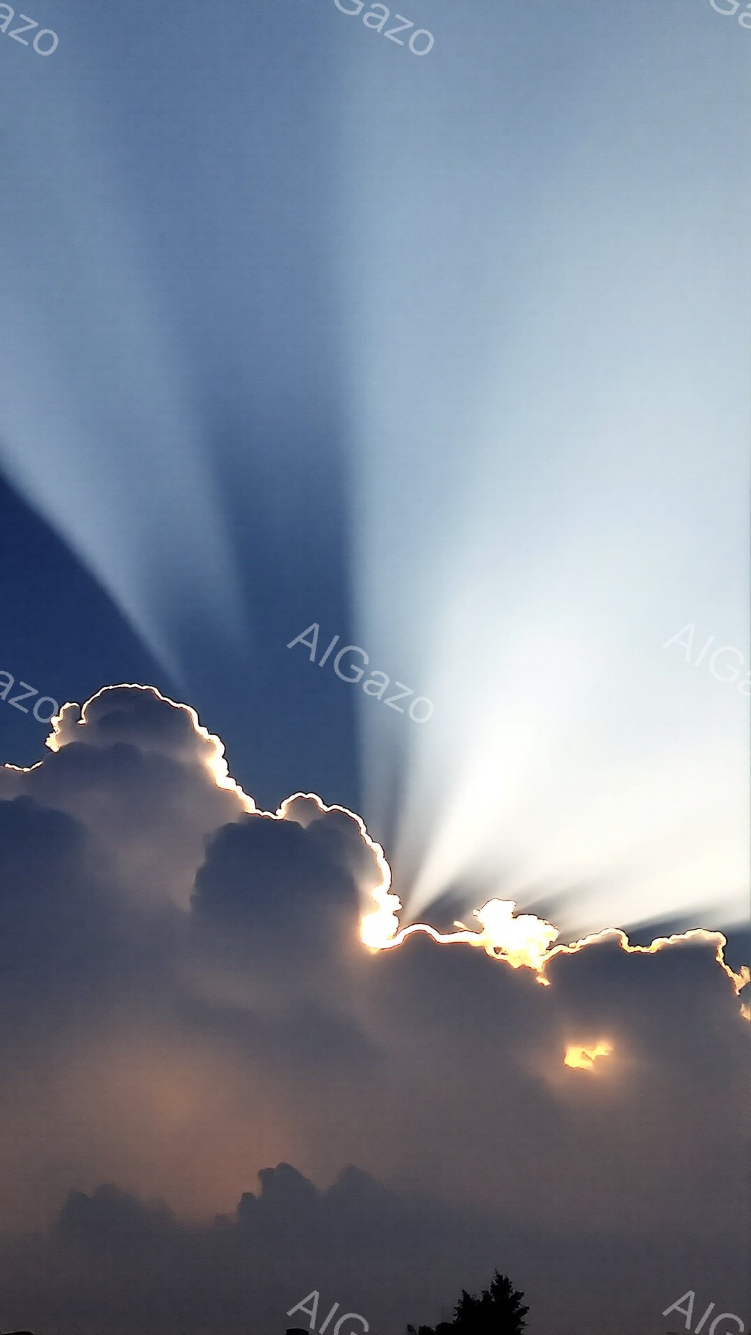 雲が広がり、その隙間から強い光線が差し込んでいる空の写真です。雲は暗いグレーと白のコントラストで、まるでドラマチックな舞台装置のようです。遠くには緑の木々が見え、夕焼けまたは日の出の穏やかで神秘的な雰囲気を醸し出しています。
