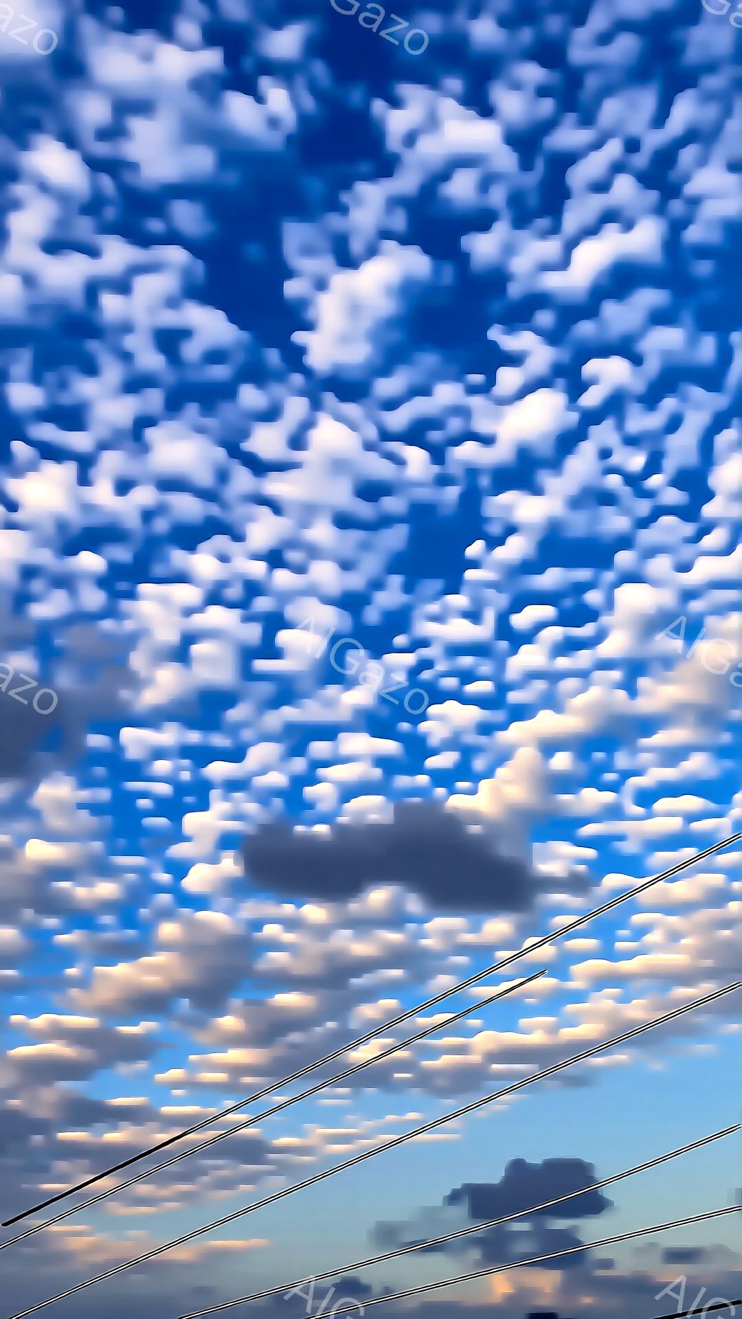 鮮やかな青空に、ふわふわとした白い雲が広がり、まるで羊雲のようです。画面下部には電線が数本走り、遠くには緑の木々がわずかに見えます。全体的に晴れやかで穏やかな、夏の日の空の雰囲気を感じさせます。