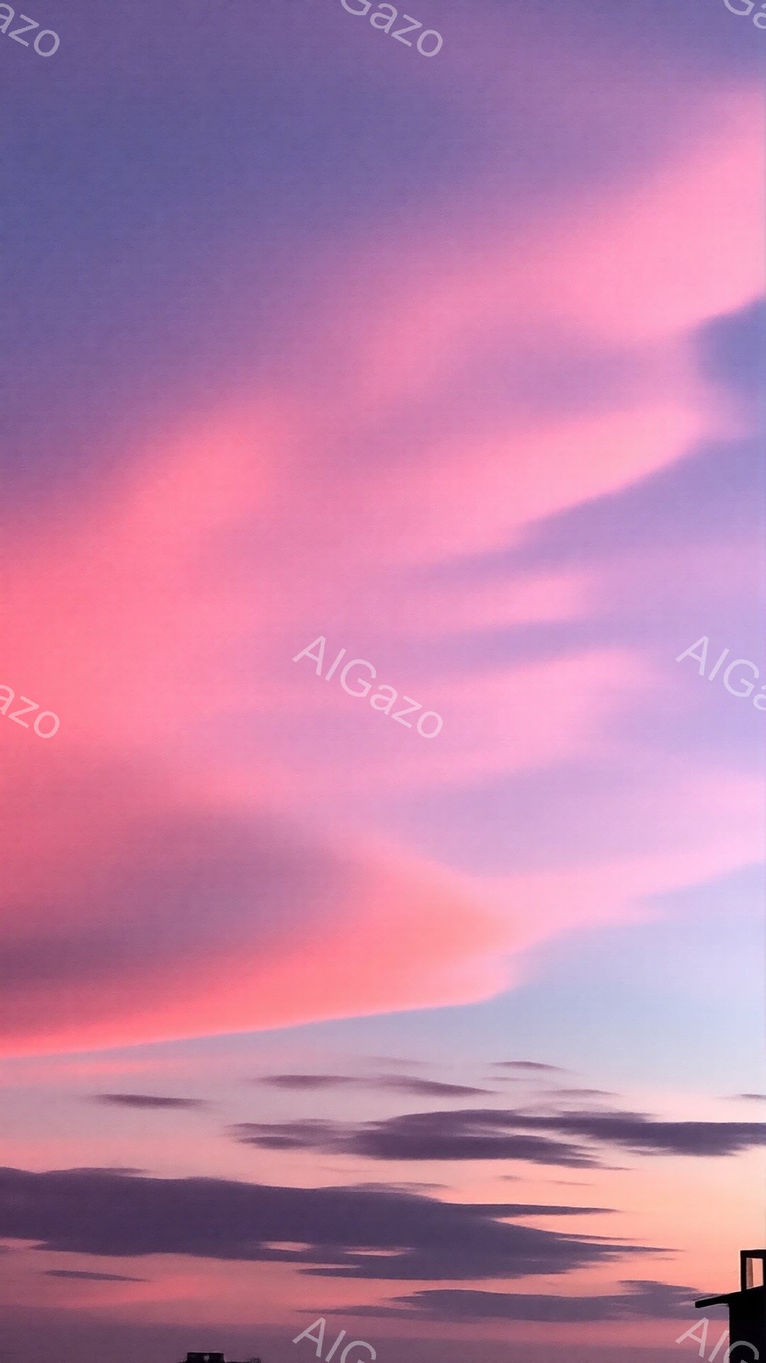 空は鮮やかなピンクと紫色の雲で覆われており、夕焼けの壮大な光景が広がっています。雲は水平方向に伸びており、まるで空を彩る帯のようです。下には、建物の一部が見え隠れし、薄暗い影がシルエットのように浮かび上がっています。