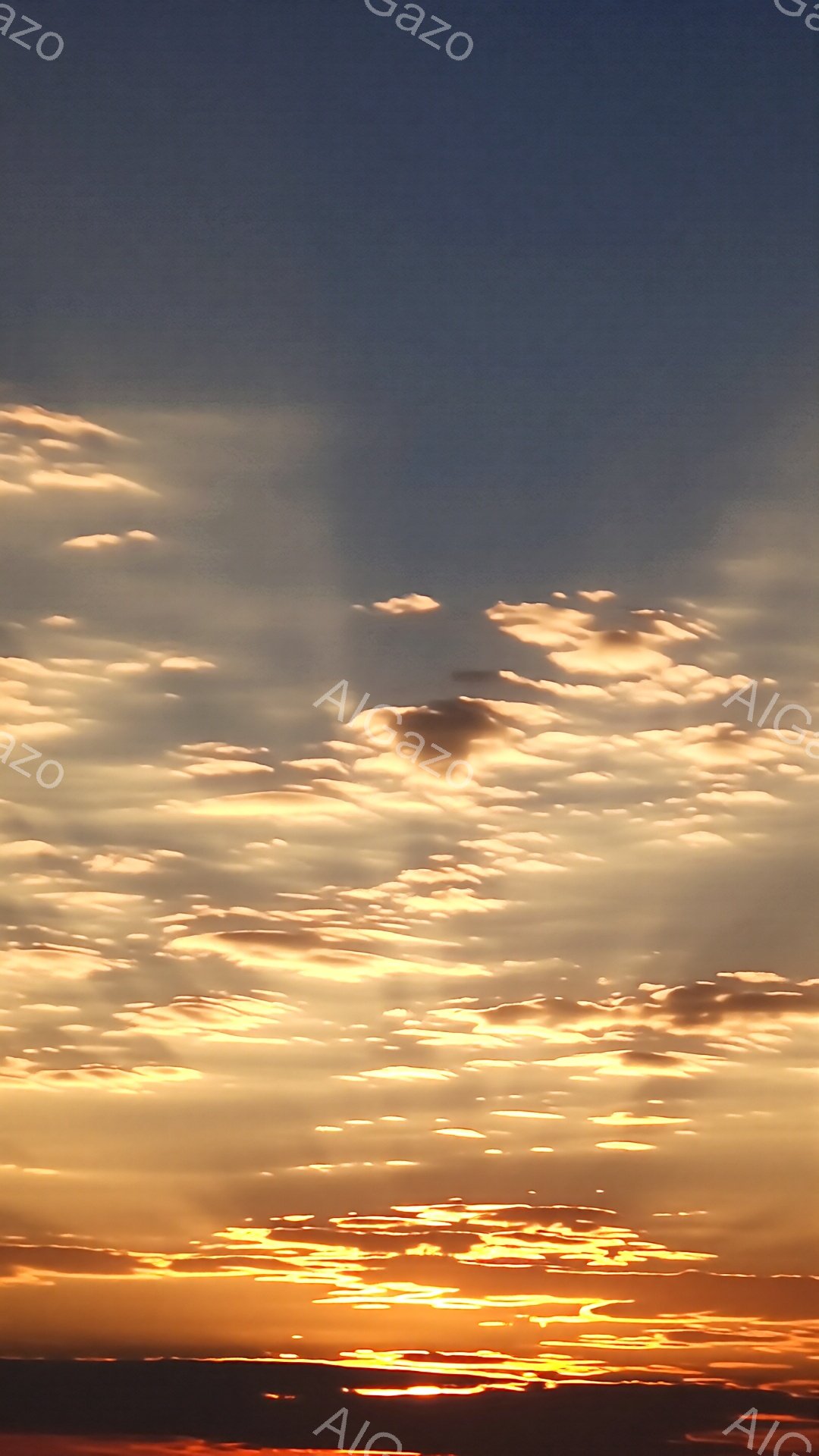 太陽が地平線に沈み、空と水面が黄金色に染まっている風景です。雲はオレンジと黄色の鮮やかな光を受けて輝き、水面には太陽の反射がキラキラと光り、まるで黄金の道が広がっているかのようです。穏やかで幻想的な雰囲気が漂い、静寂と安らぎを感じさせます。