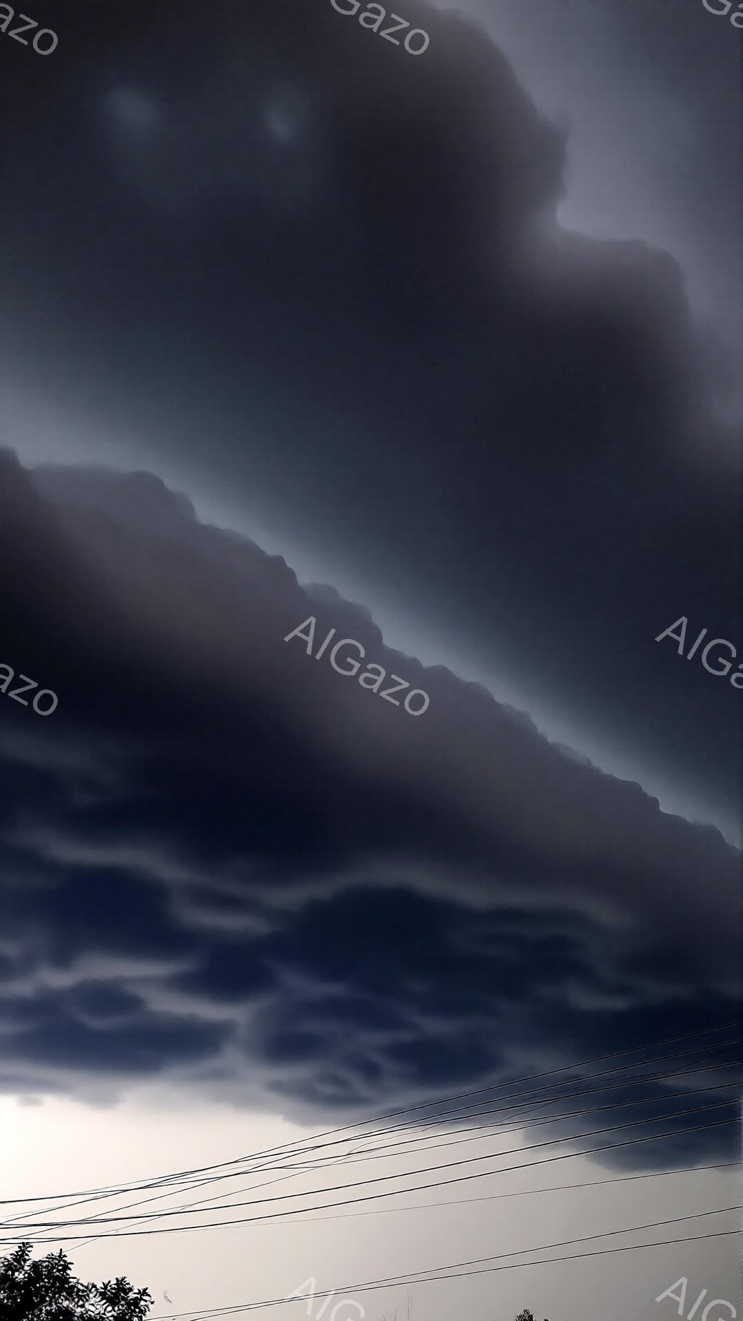 濃い灰色の雲が空を覆い、まるで嵐が近づいているかのような重々しい雰囲気です。雲は独特の形をしており、まるでうねる波のようです。地平線には木々の緑と電線がわずかに見え、迫り来る雲とのコントラストが印象的です。