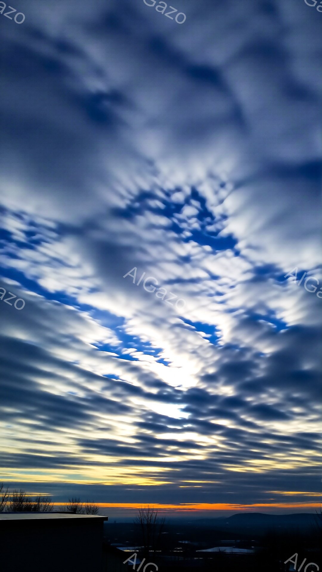 雲海が広がり、地平線にはオレンジ色の光が差し込んでいます。空には規則的に並んだ雲が特徴的で、まるで波紋のような模様を形作っています。画面下部には、木の枝がシルエットとして写り込み、冬の寒々とした風景を暗示しています。